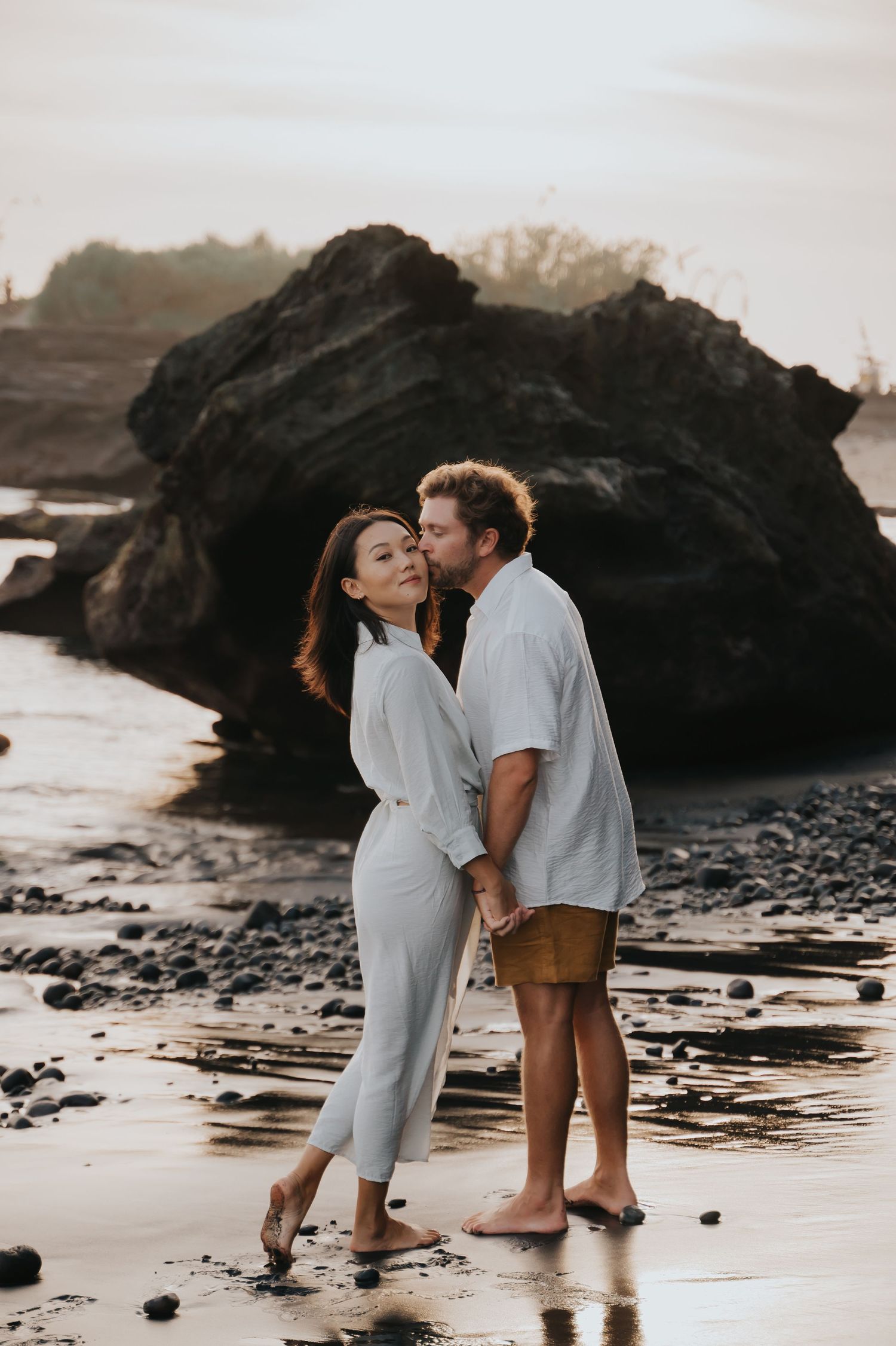 A couple shares an intimate moment on a rocky beach at sunset, standing barefoot in the wet sand near large boulders.