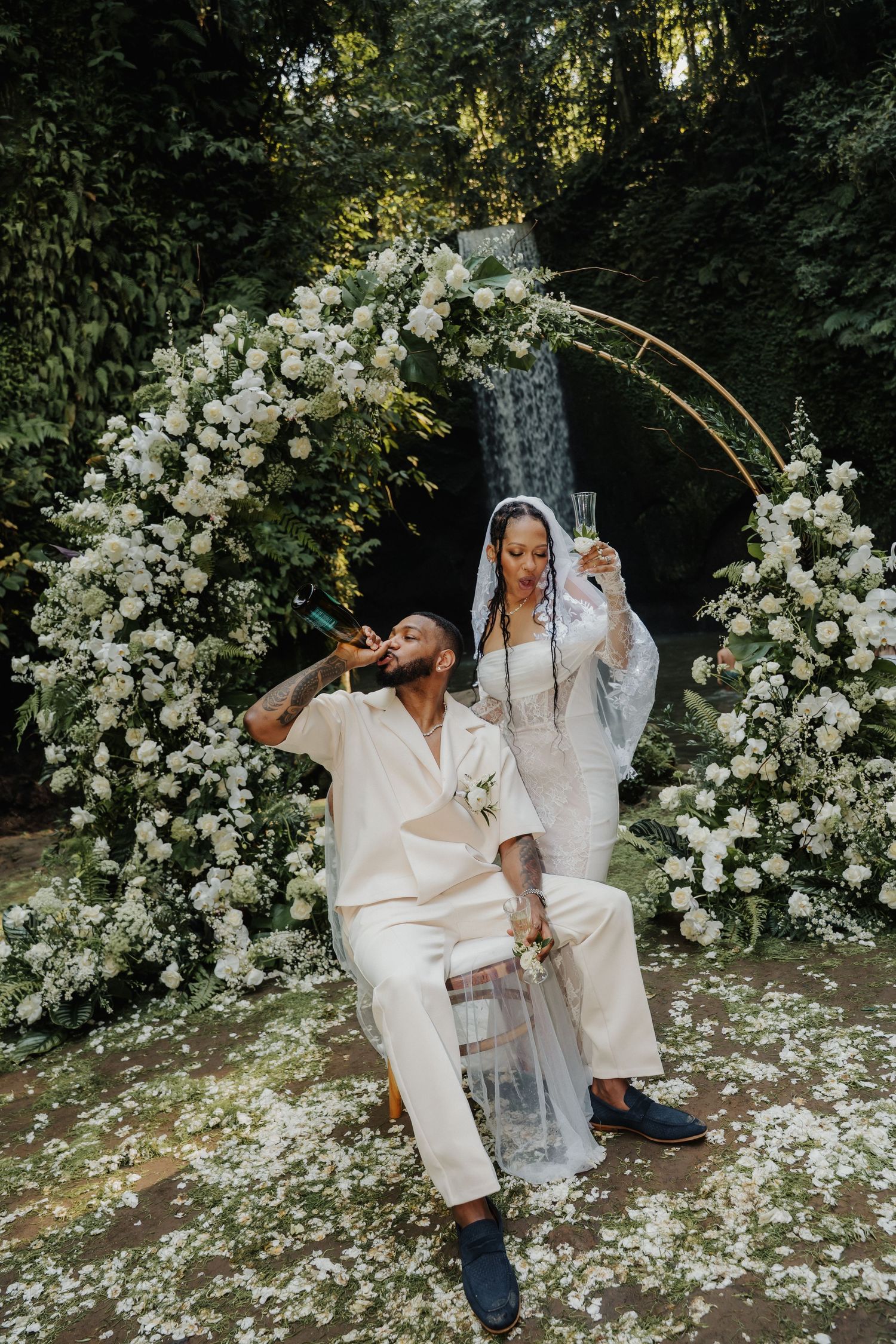 Newlyweds celebrate with champagne under a floral arch with waterfall backdrop in matching ivory outfits.