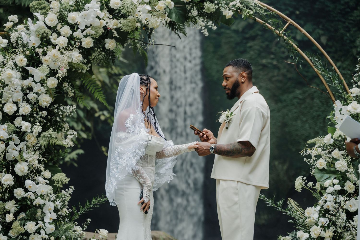 Groom placing ring on bride's finger under white flower arch at tropical waterfall wedding.