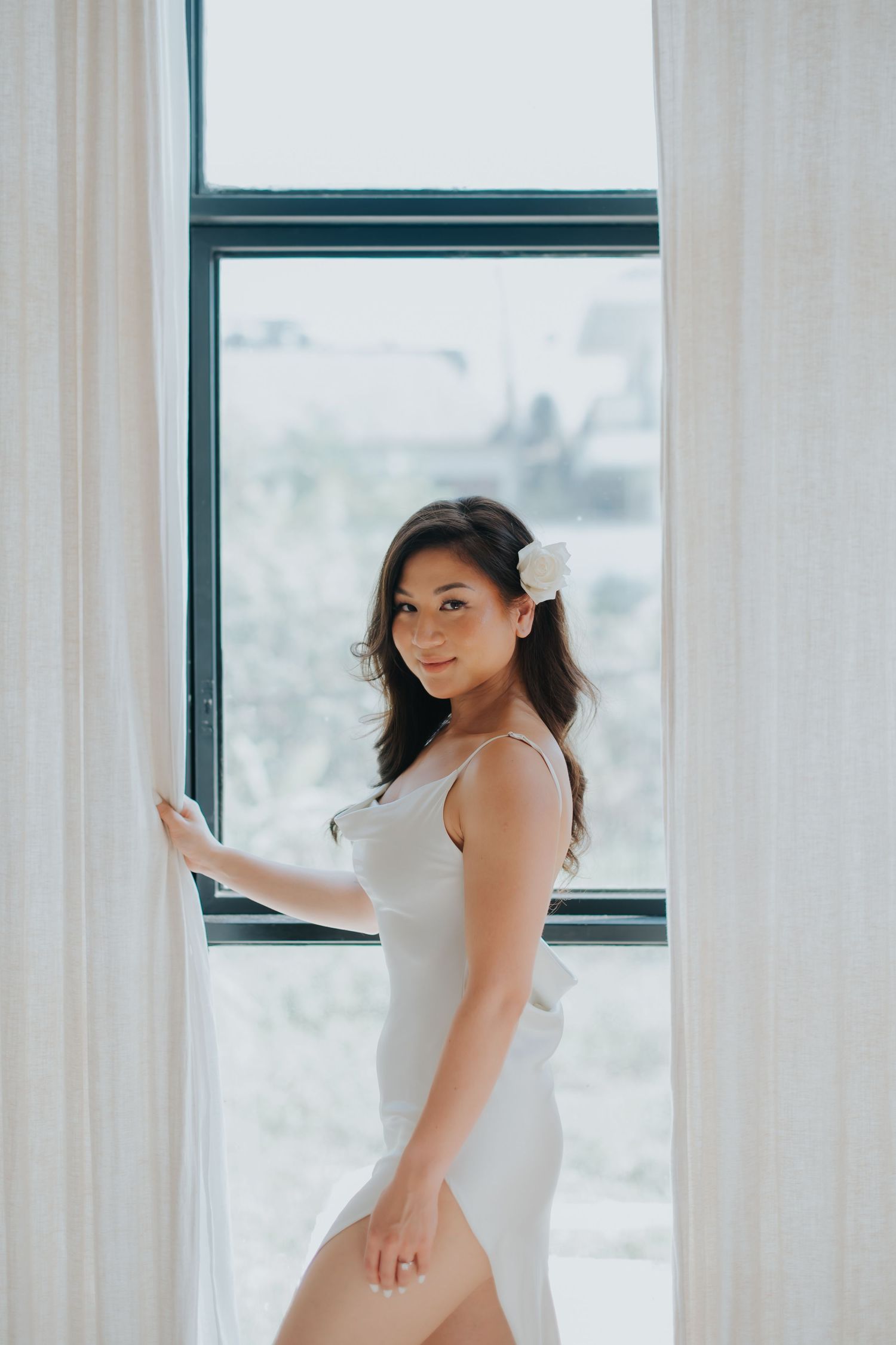 Young Asian woman in white dress with flower hair accessory stands by bright window holding sheer curtain.