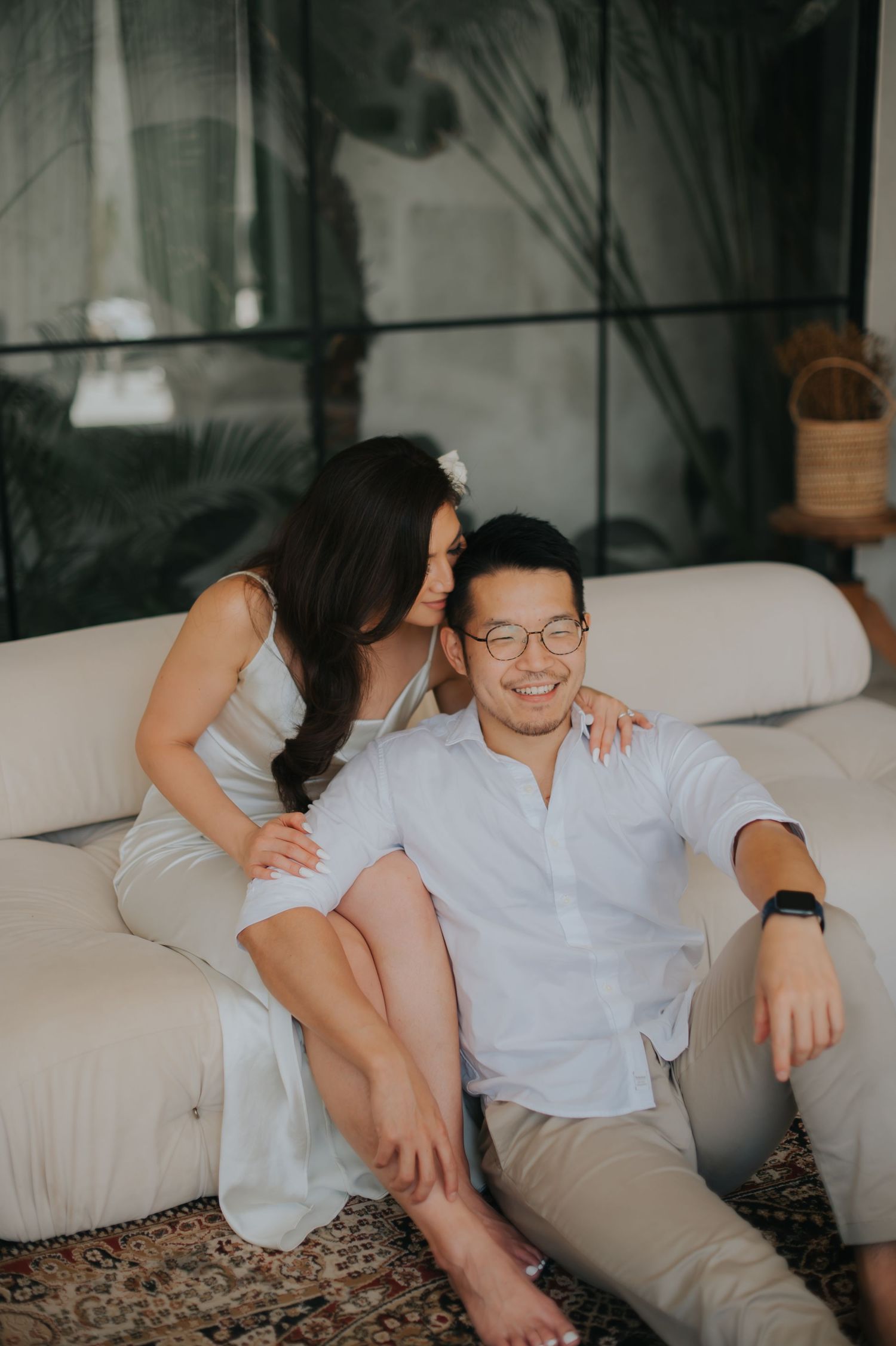 A couple in white outfits share a tender moment on a cream sofa, woman embracing man from behind as he smiles warmly.