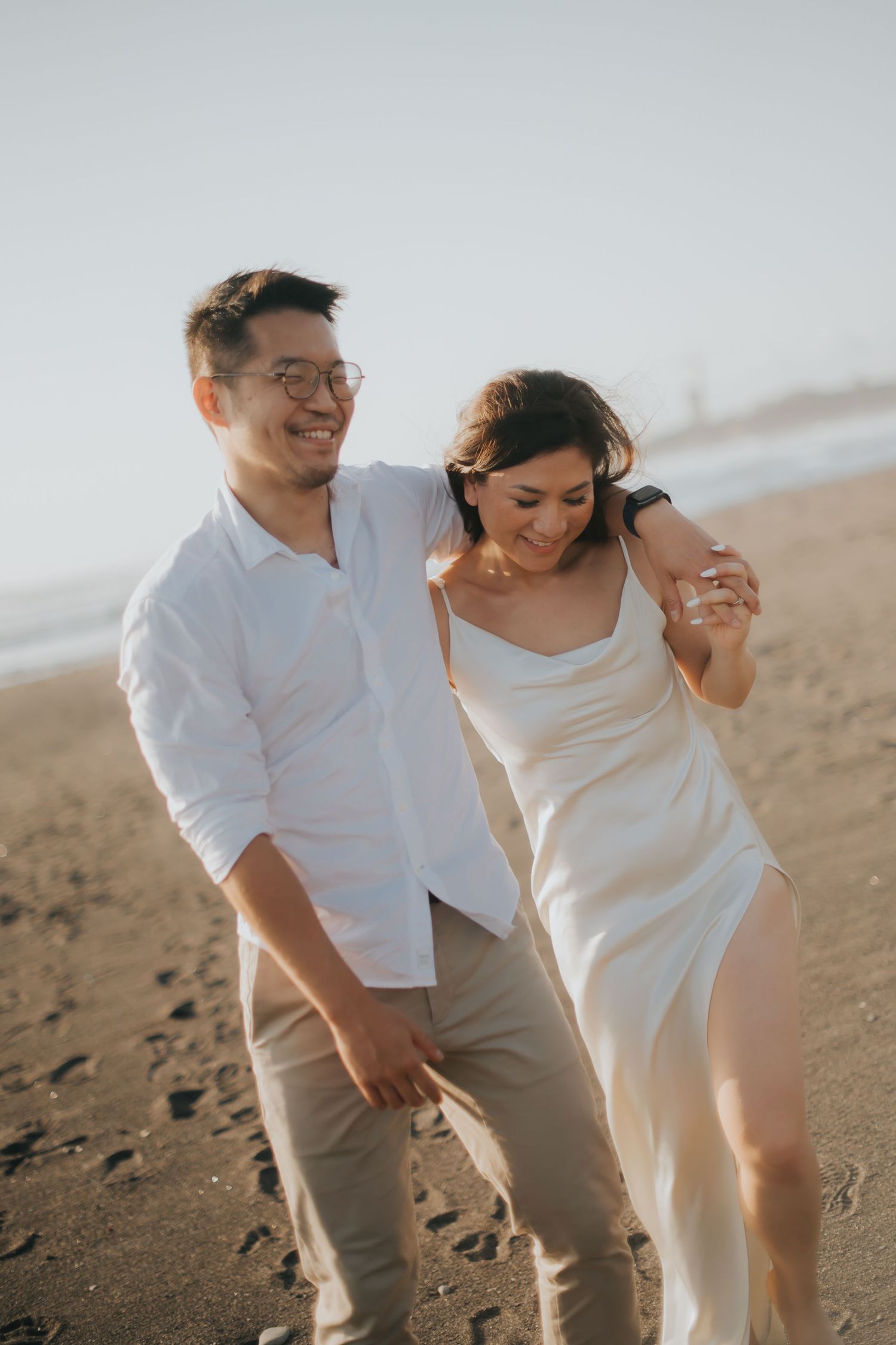 Couple walking on beach, man in white shirt and glasses, woman in white dress, smiling together at sunset.