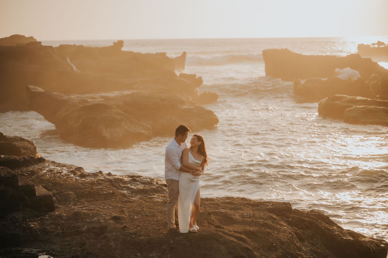 Couple embracing on rocky ocean shore at golden sunset, waves and dramatic rock formations in background.