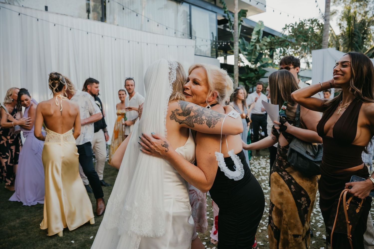 Bride in satin gown and veil hugs tattooed guest at joyful outdoor tropical wedding reception celebration.