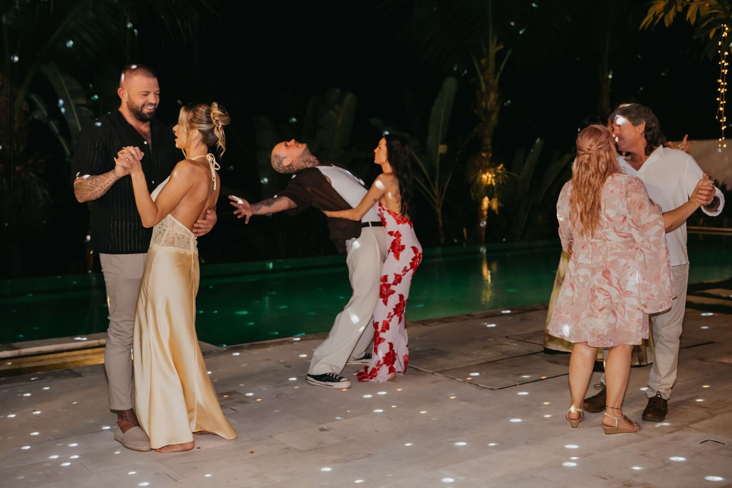 Couples dance poolside at night during outdoor wedding reception surrounded by tropical greenery.