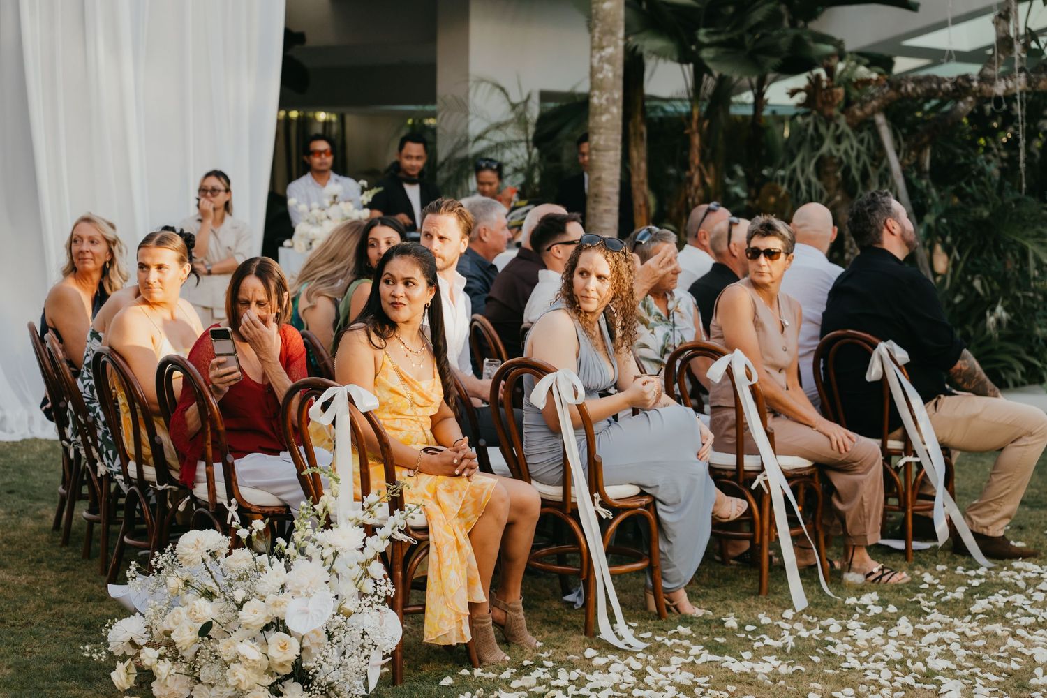 Wedding guests seated in wooden chairs at an outdoor tropical ceremony surrounded by white floral arrangements.