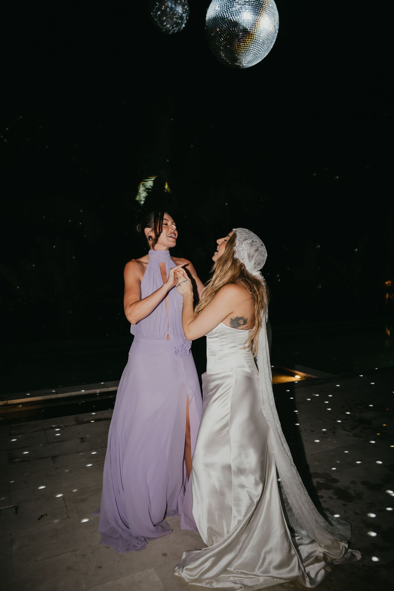 Bride in white satin gown and bridesmaid in lavender dress dancing under disco balls at night.