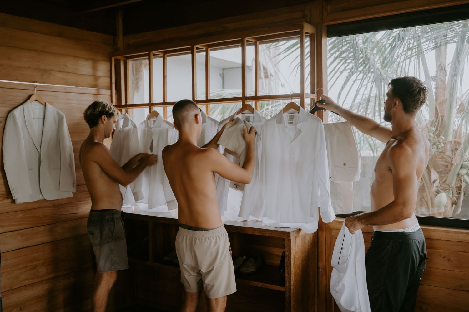 Three shirtless groomsmen selecting white dress shirts from hangers in a wooden tropical wedding prep room.