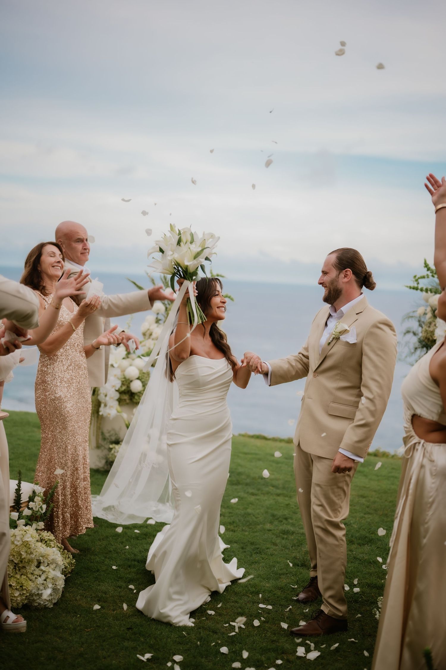 Newlyweds walk down the aisle as guests toss white flower petals at their outdoor oceanview wedding ceremony.
