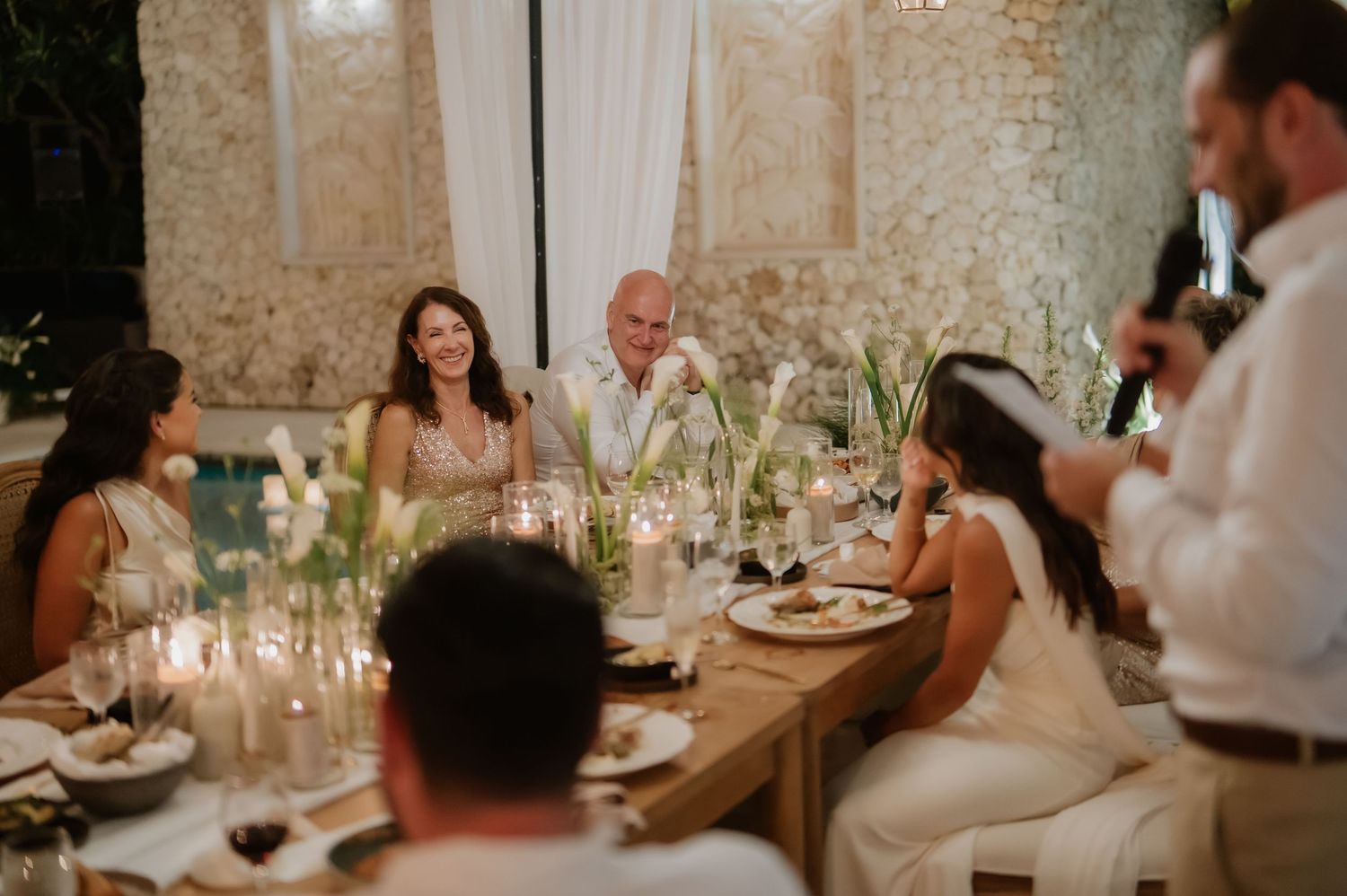 Wedding guests laugh and converse at candlelit reception dinner table with elegant white floral decor.