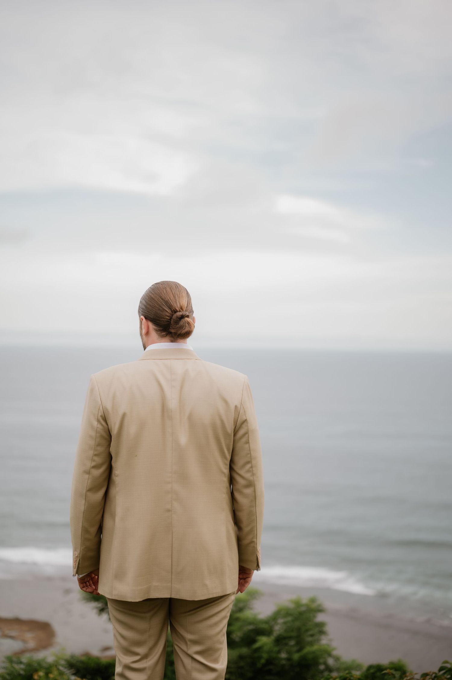 Man in beige suit with hair bun gazes at ocean view on overcast day.