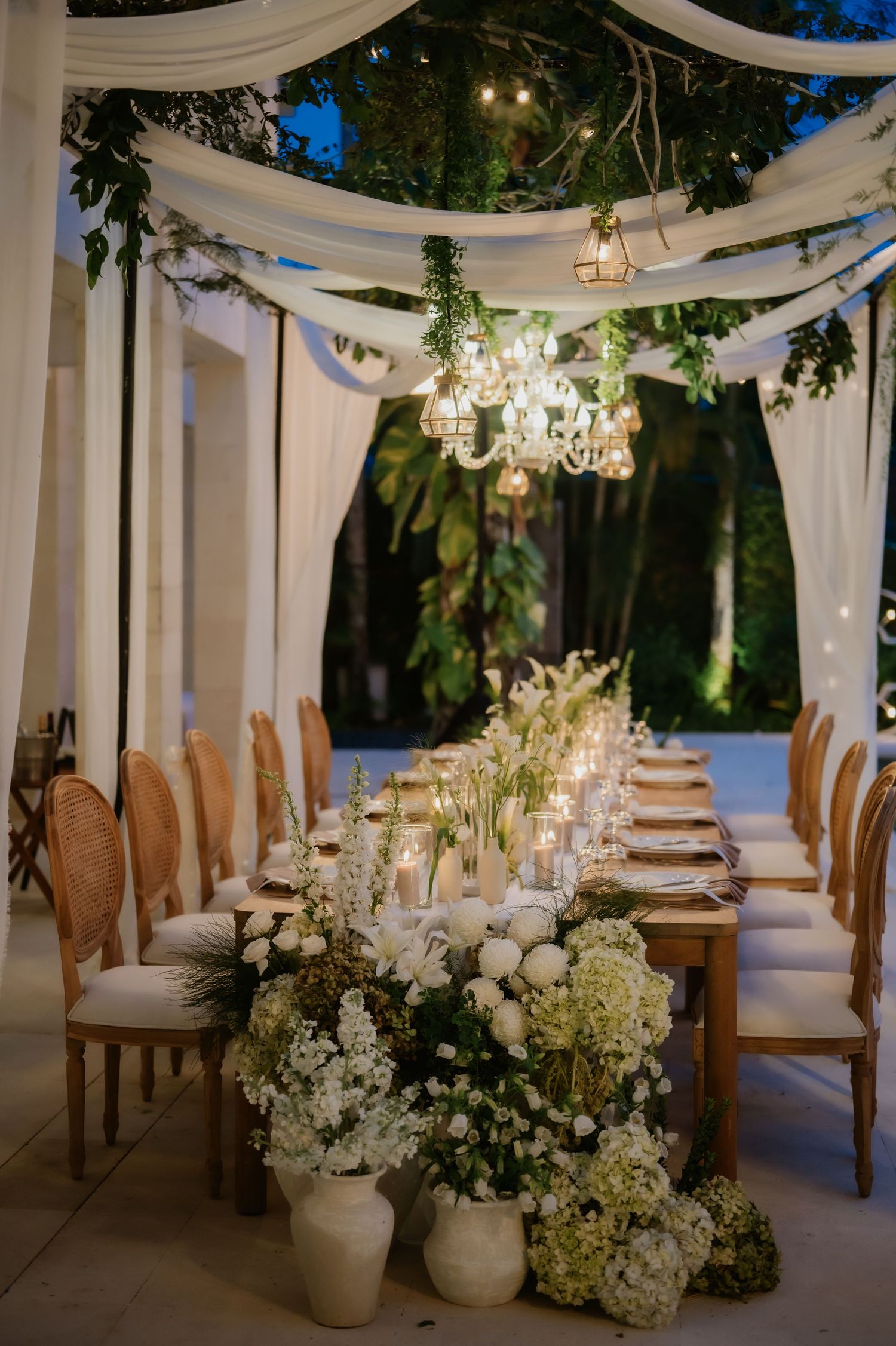 Romantic outdoor wedding dinner table with white florals, candles, and crystal chandeliers at dusk.