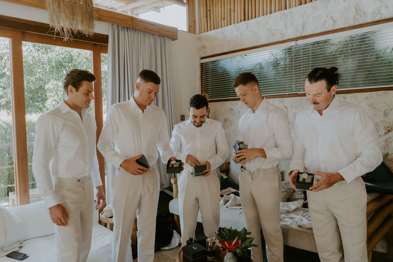 Groomsmen in white shirts and tan pants share drinks together in a bright tropical villa room.
