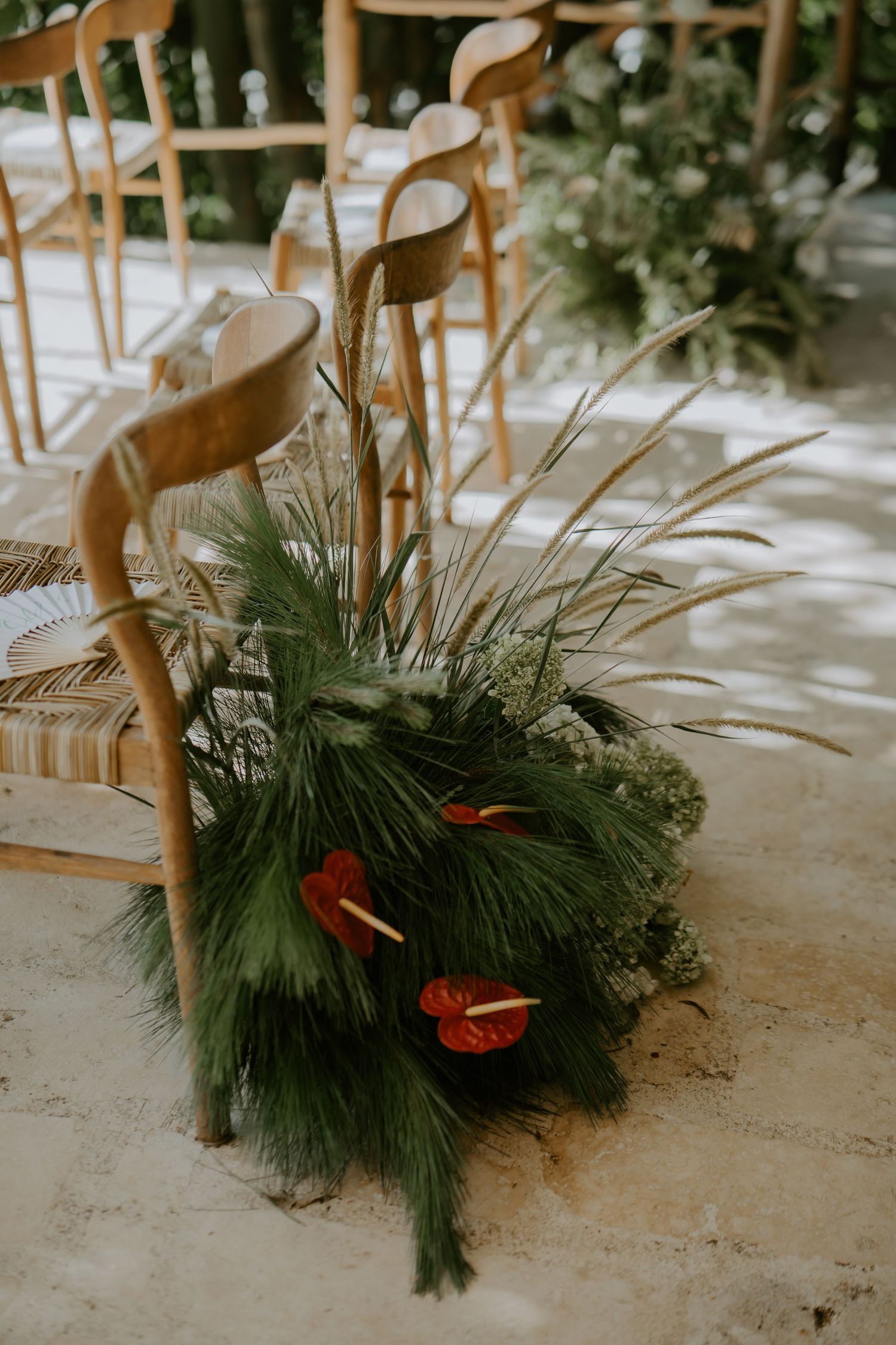 Wedding aisle floral arrangement featuring red anthuriums, pine, and pampas grass on stone floor beside wooden chair.