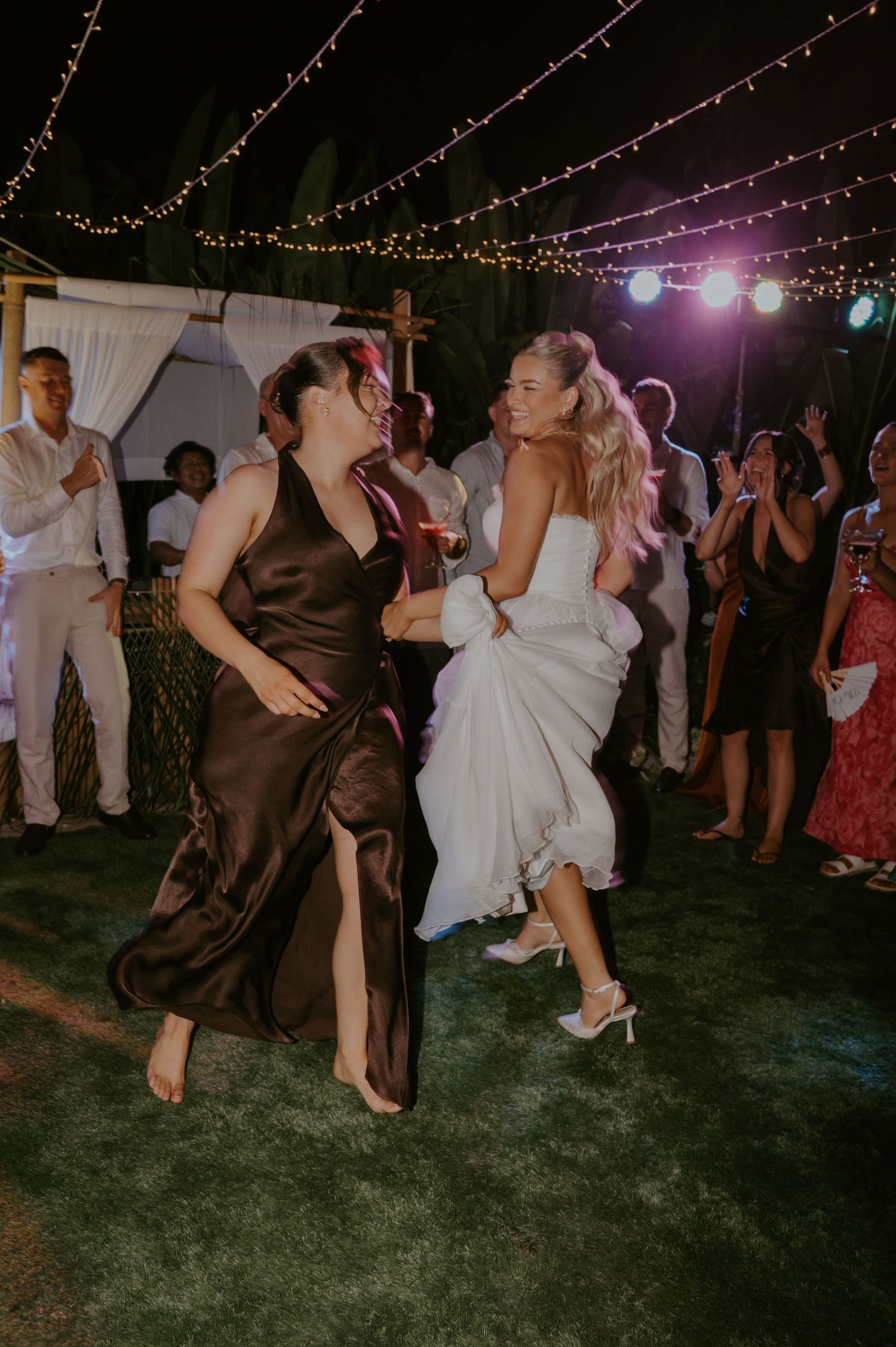Bride in white gown dancing joyfully with bridesmaid in brown dress at outdoor wedding reception.
