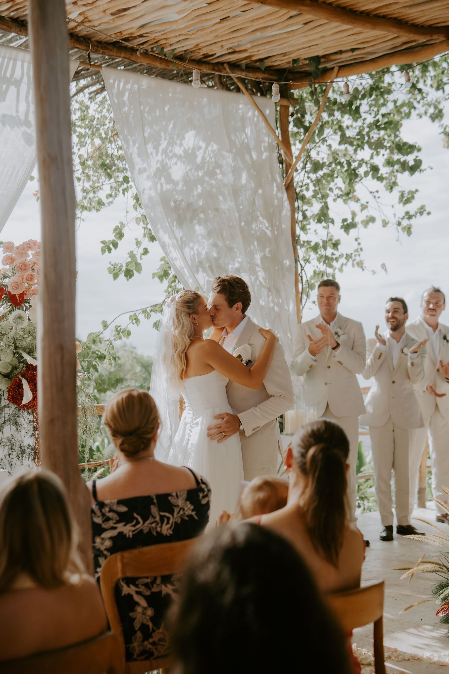 Bride and groom share first kiss at outdoor tropical wedding ceremony with guests applauding.