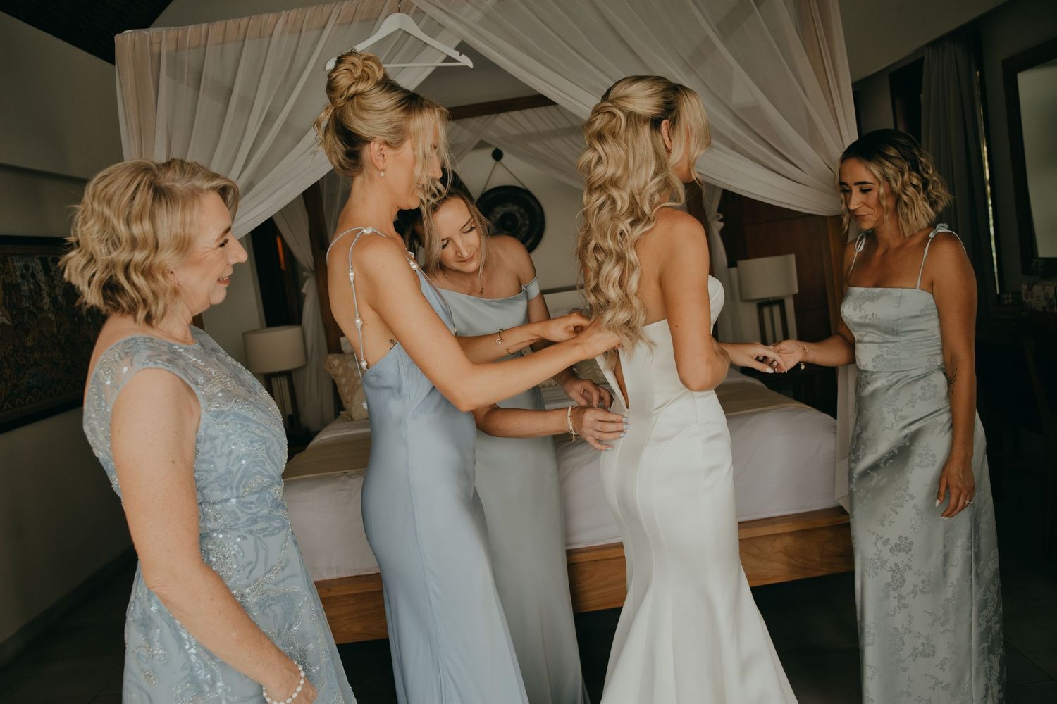 Bride in white gown surrounded by bridesmaids in blue dresses during wedding preparations.