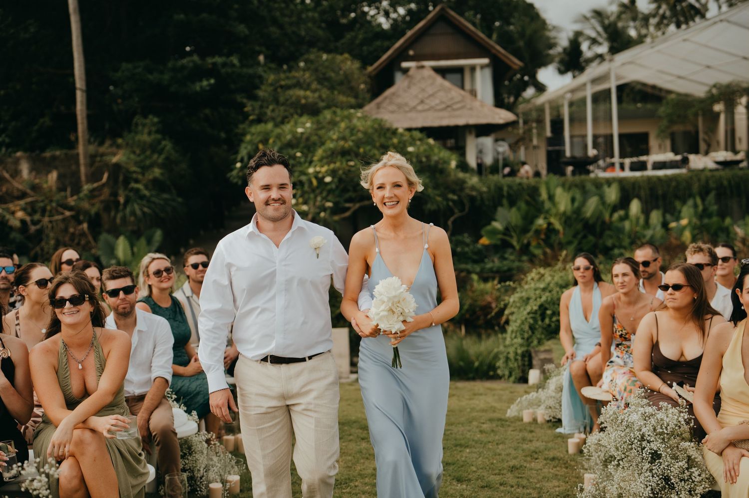 Smiling bridesmaid in blue gown and groomsman walk down aisle past seated wedding guests outdoors.