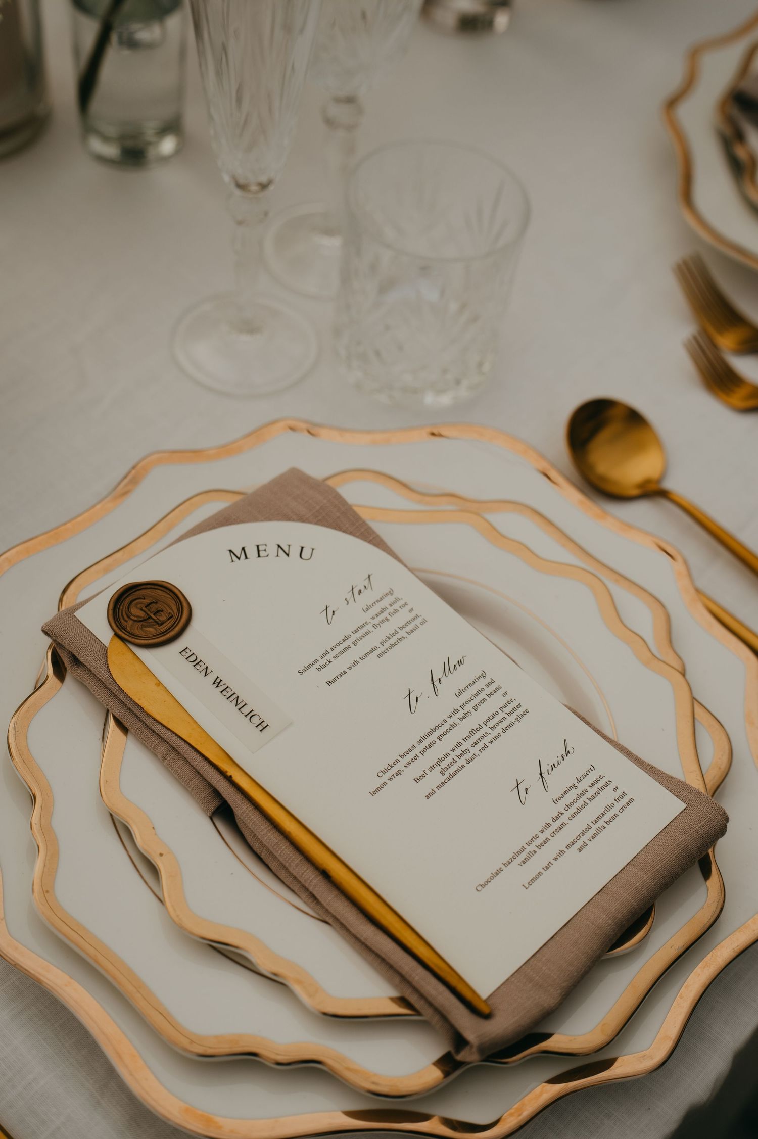 Elegant wedding place setting with gold-rimmed plates, wax seal menu card, and gold cutlery on white linen.