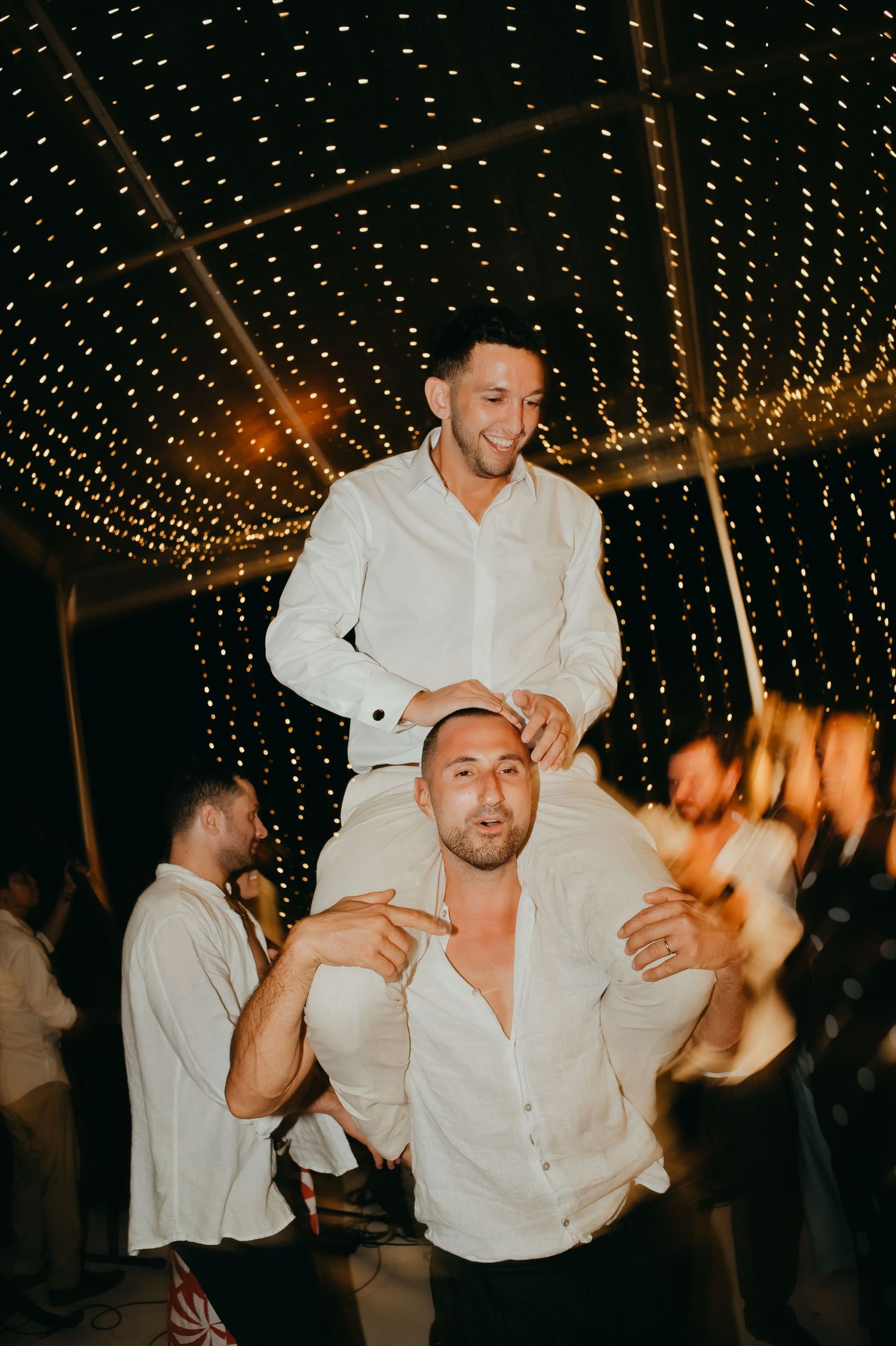 Groomsman lifted on shoulders celebrating under a canopy of twinkling fairy lights at a wedding reception.