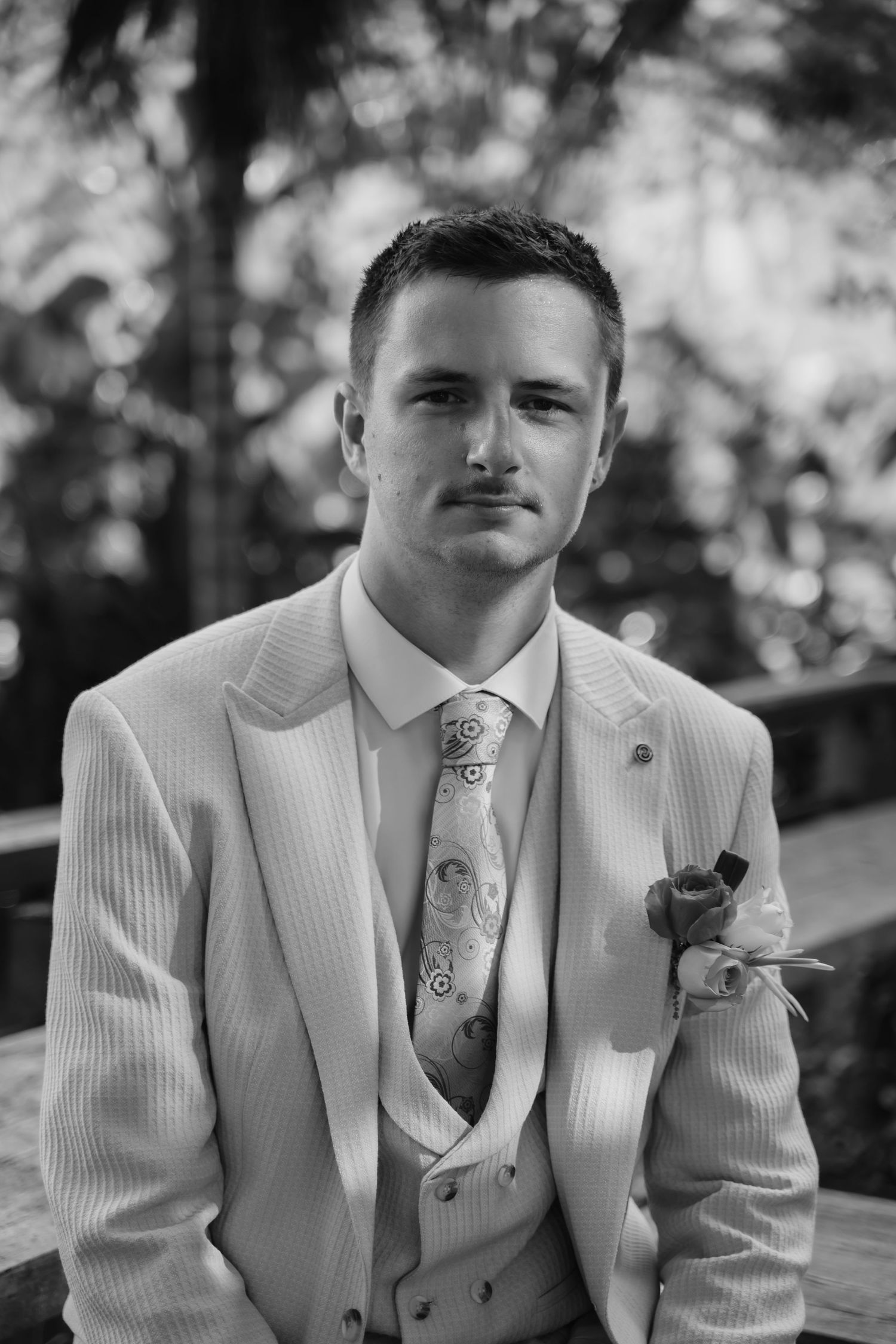Groom in light suit and floral tie with boutonniere posing outdoors in black and white portrait.