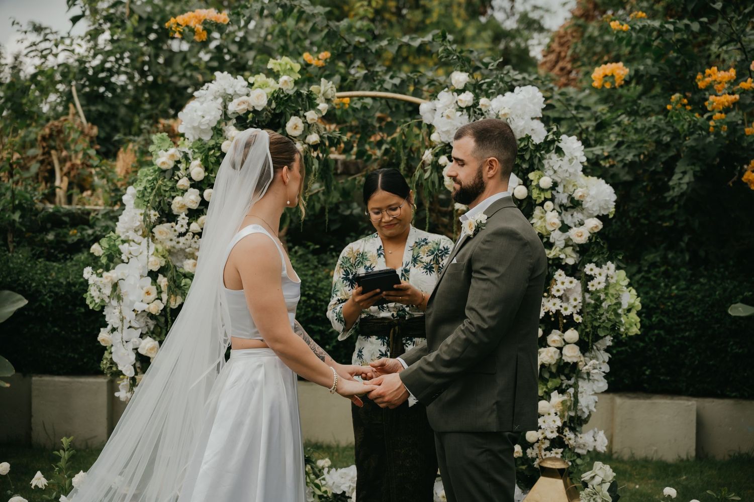 Bride and groom exchange vows under white floral arch with officiant in lush garden wedding ceremony.