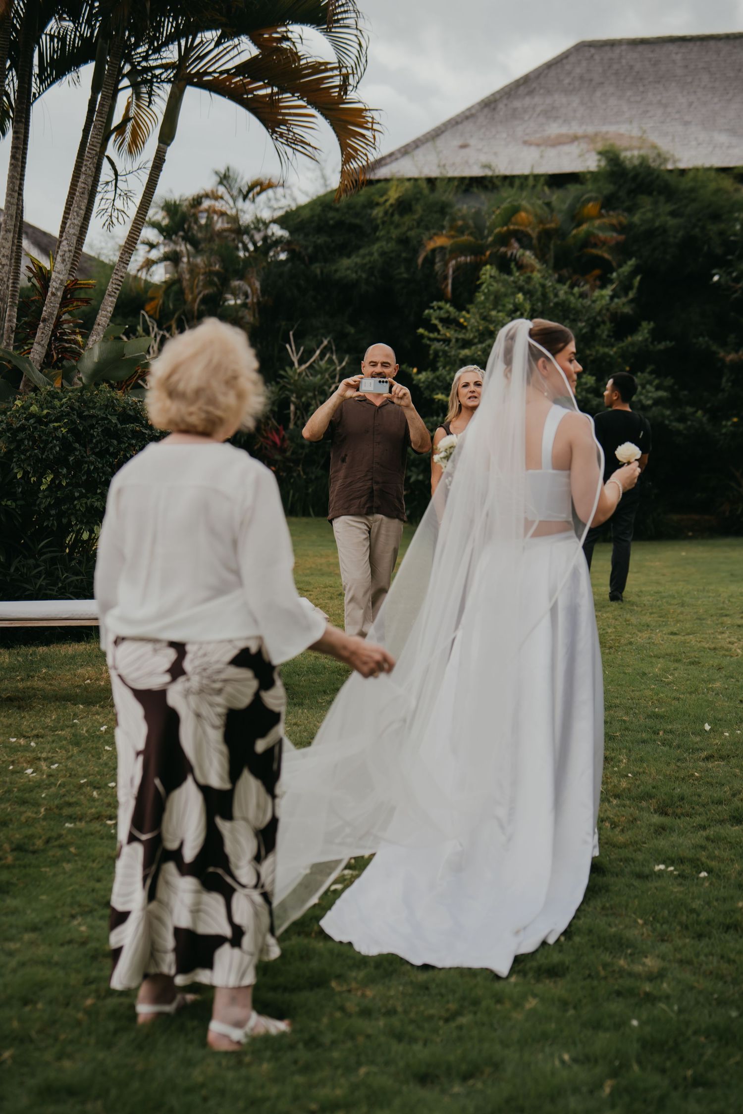 Wedding guests photograph bride in white gown and veil on tropical garden lawn.