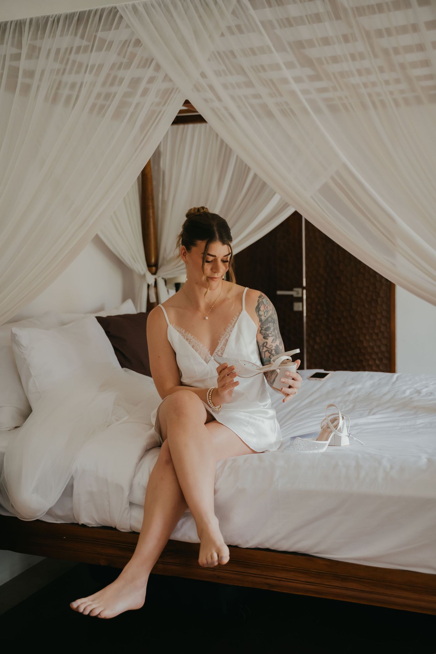 Bride in white lace slip dress sits on canopy bed holding wedding shoes in tropical villa.