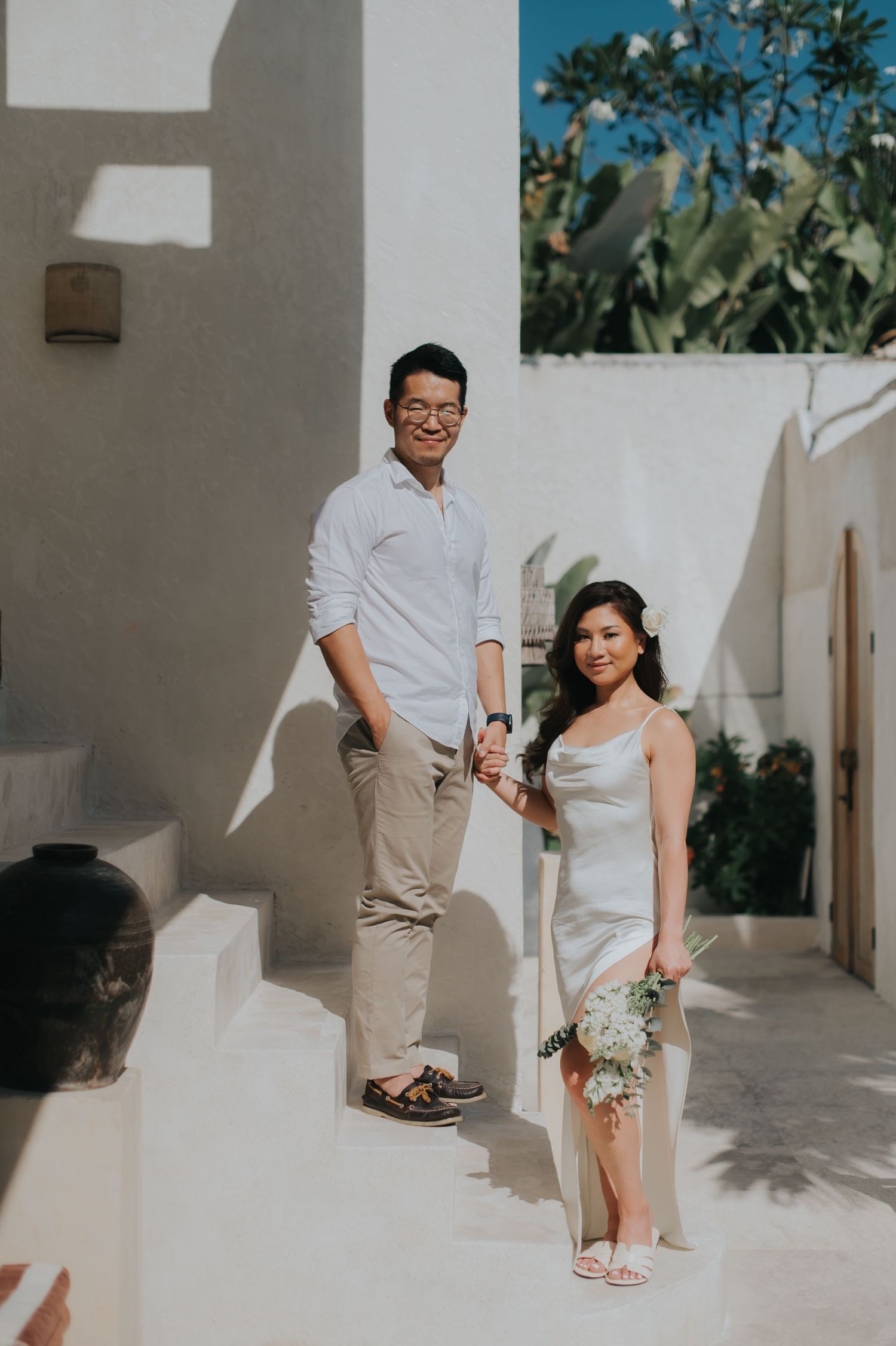 Couple in white attire holding hands on white steps, woman holding bouquet, at a sunny outdoor wedding venue.