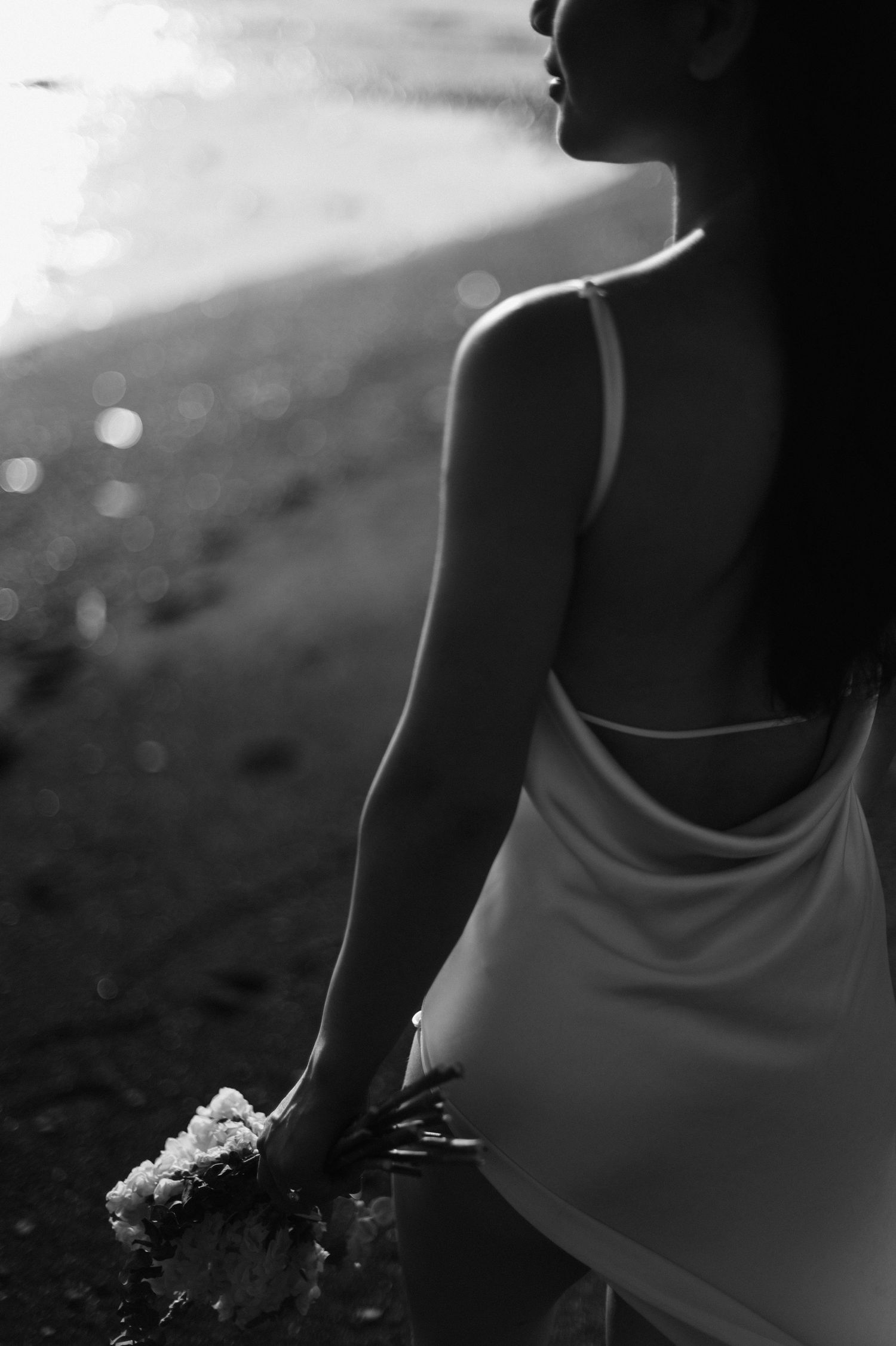 Black and white photo of a woman in a white dress holding flowers by a sunlit beach.