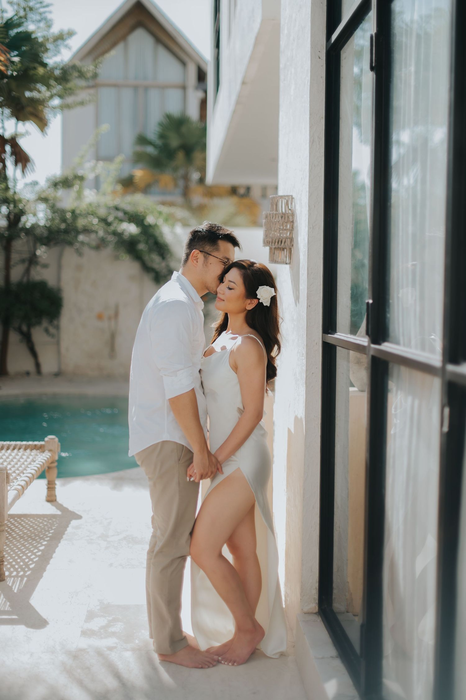 Couple sharing a romantic moment by a pool, man in white shirt kissing woman in elegant white dress at a luxury villa.