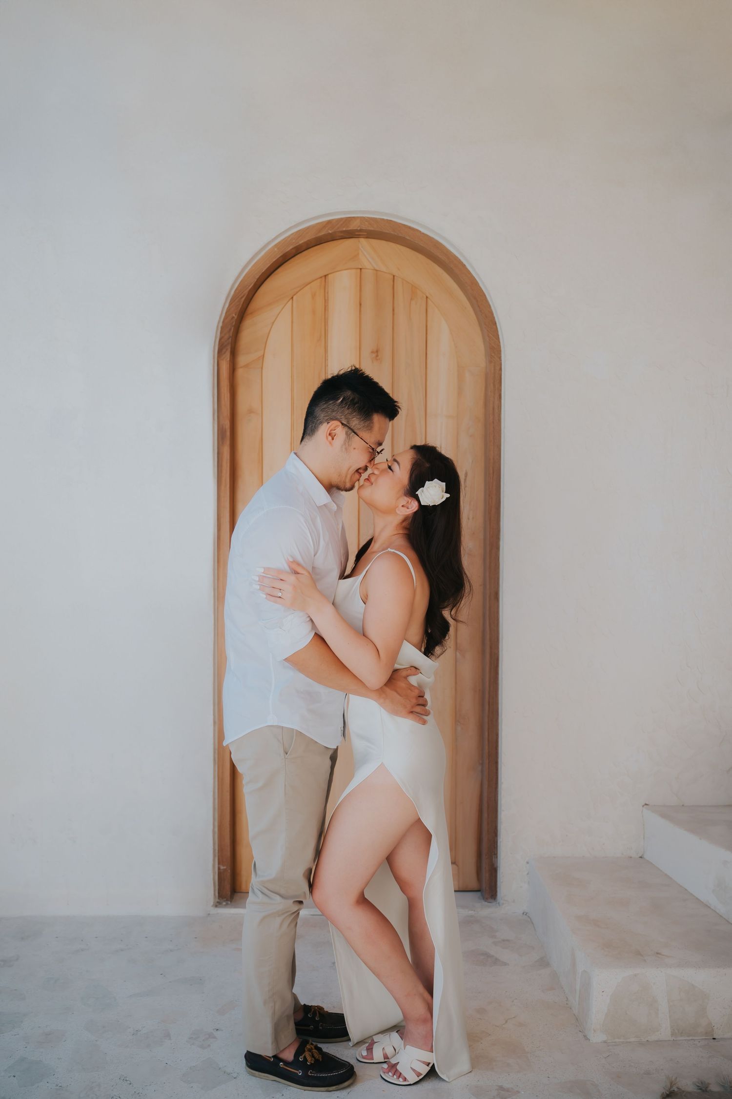 Couple sharing a romantic kiss in front of an arched wooden door, dressed in white outfits for an engagement photo.