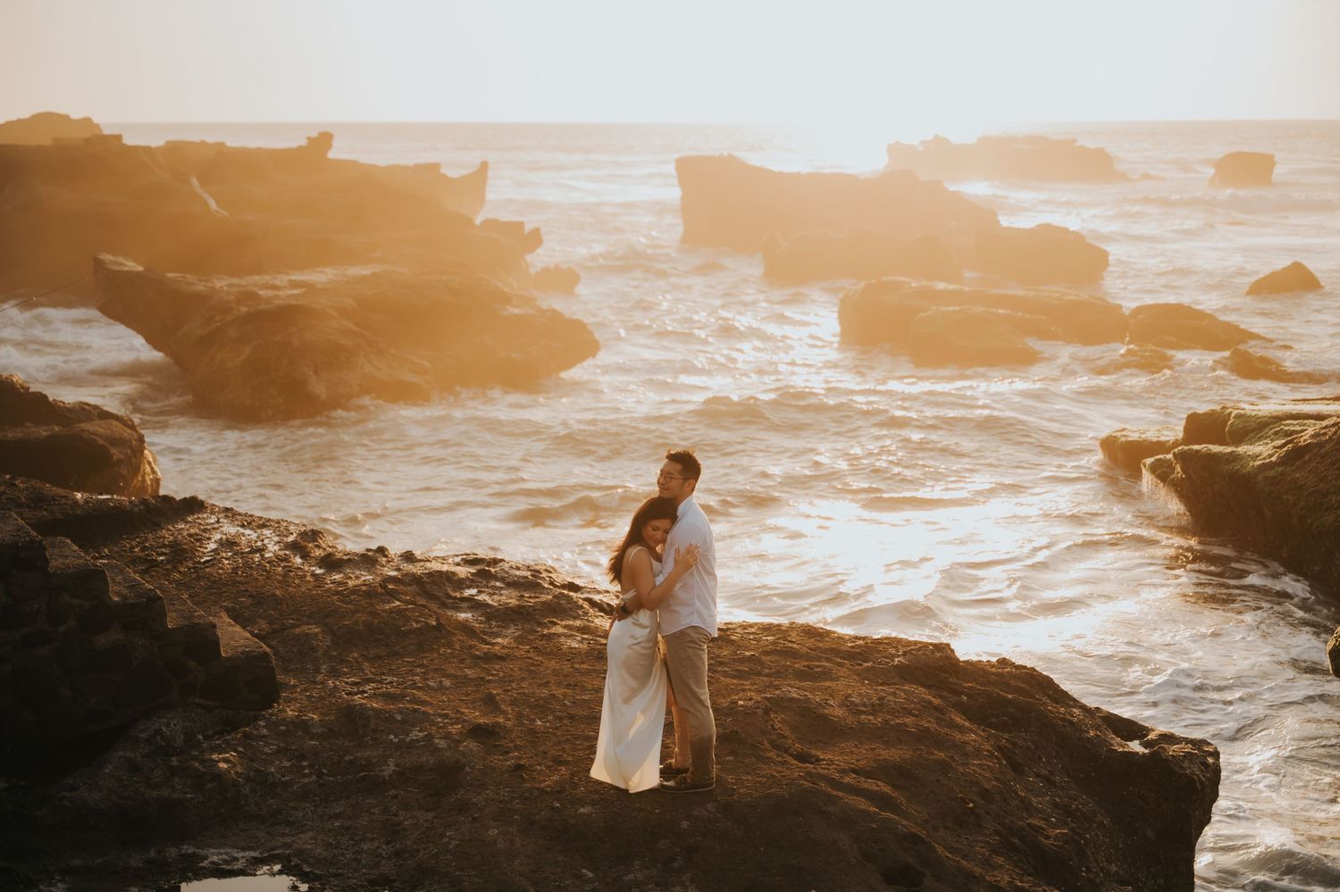 Couple embracing on rocky coastal cliffs at golden sunset with waves crashing behind them.