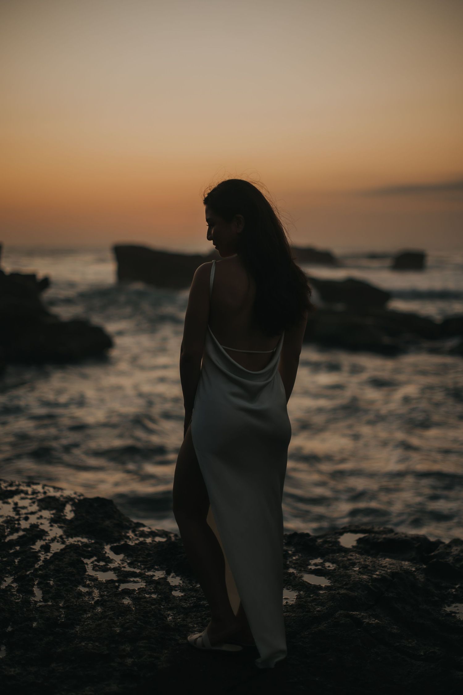 Woman in white backless dress standing on rocky shore at sunset with ocean waves in background.