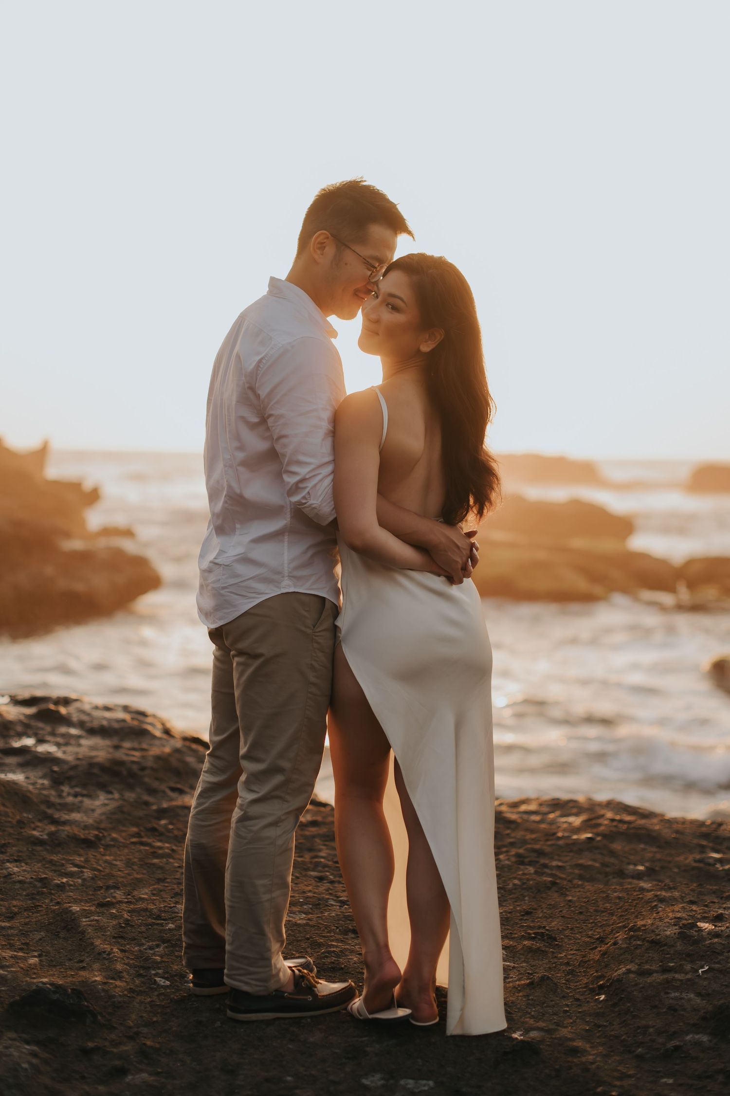 Couple embracing at sunset on rocky beach, woman in white slit dress, man in white shirt, golden hour lighting.