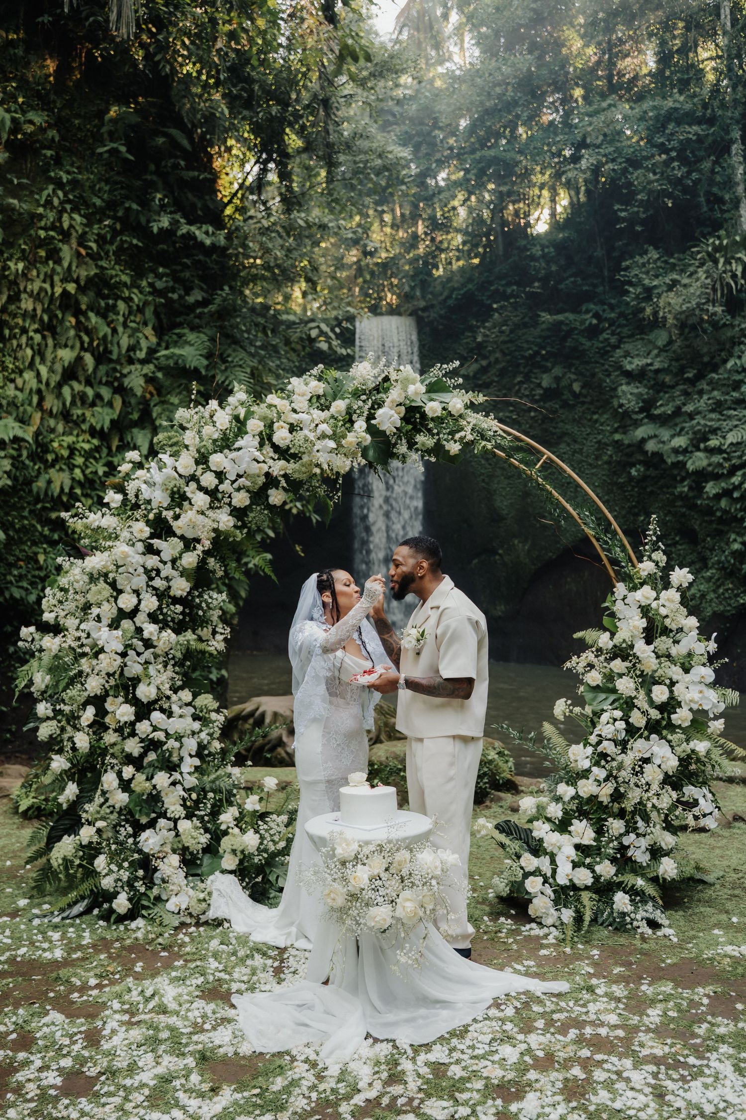 Bride and groom share cake beneath a circular white floral arch with waterfall in background.
