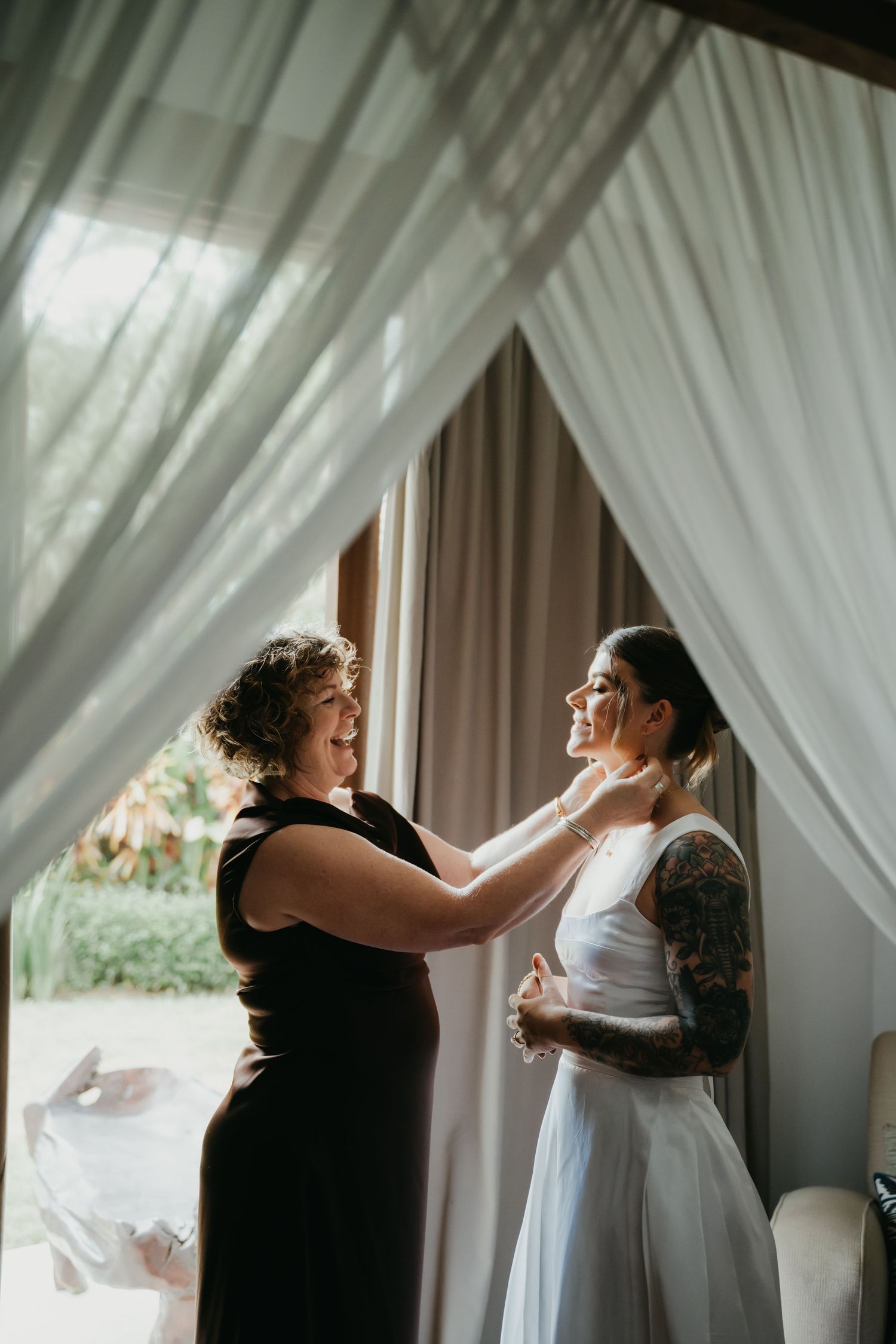 Woman in black dress adjusts bride's necklace near sheer curtains in soft natural light.