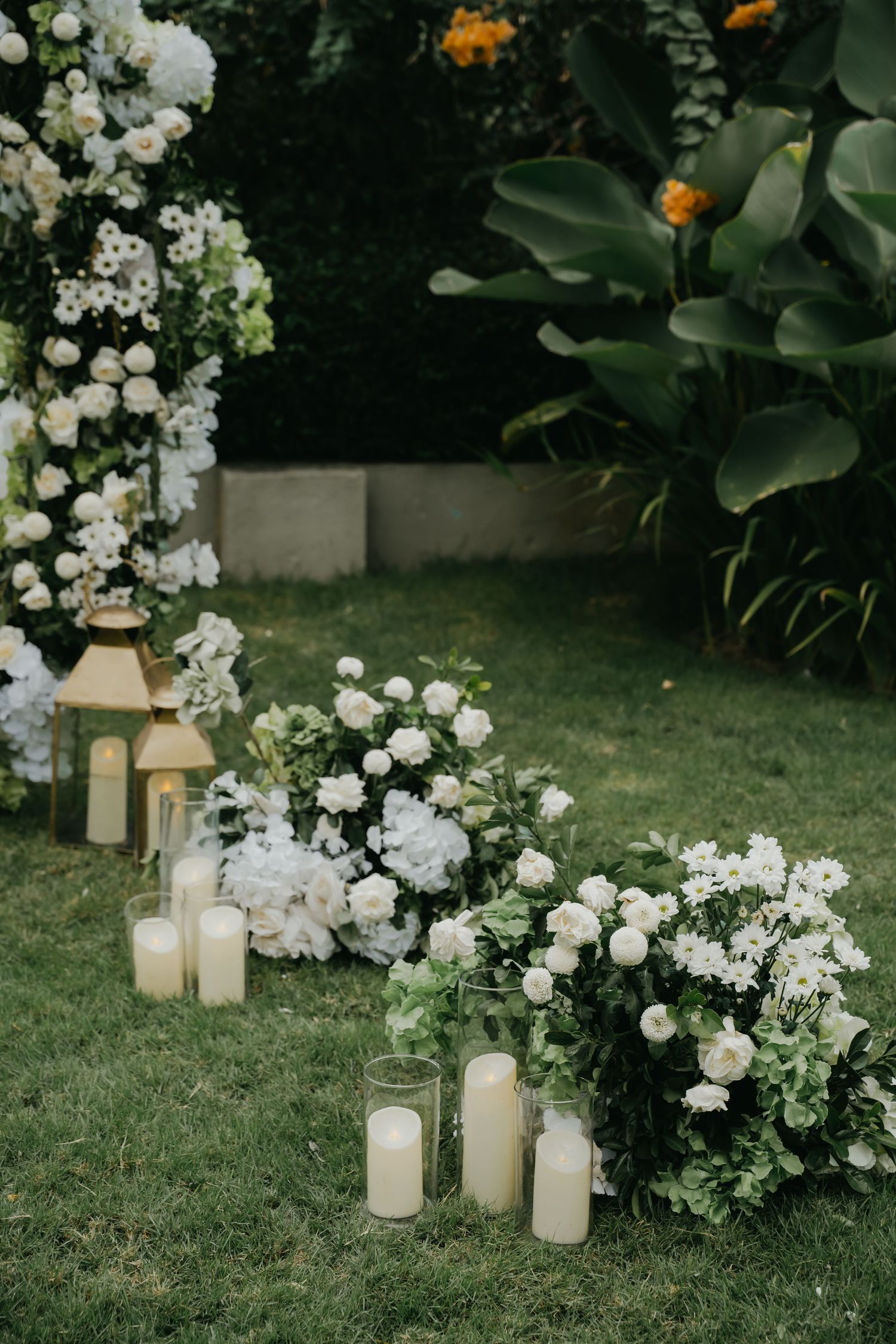White floral arrangements with hydrangeas and roses beside pillar candles on green lawn for outdoor wedding.