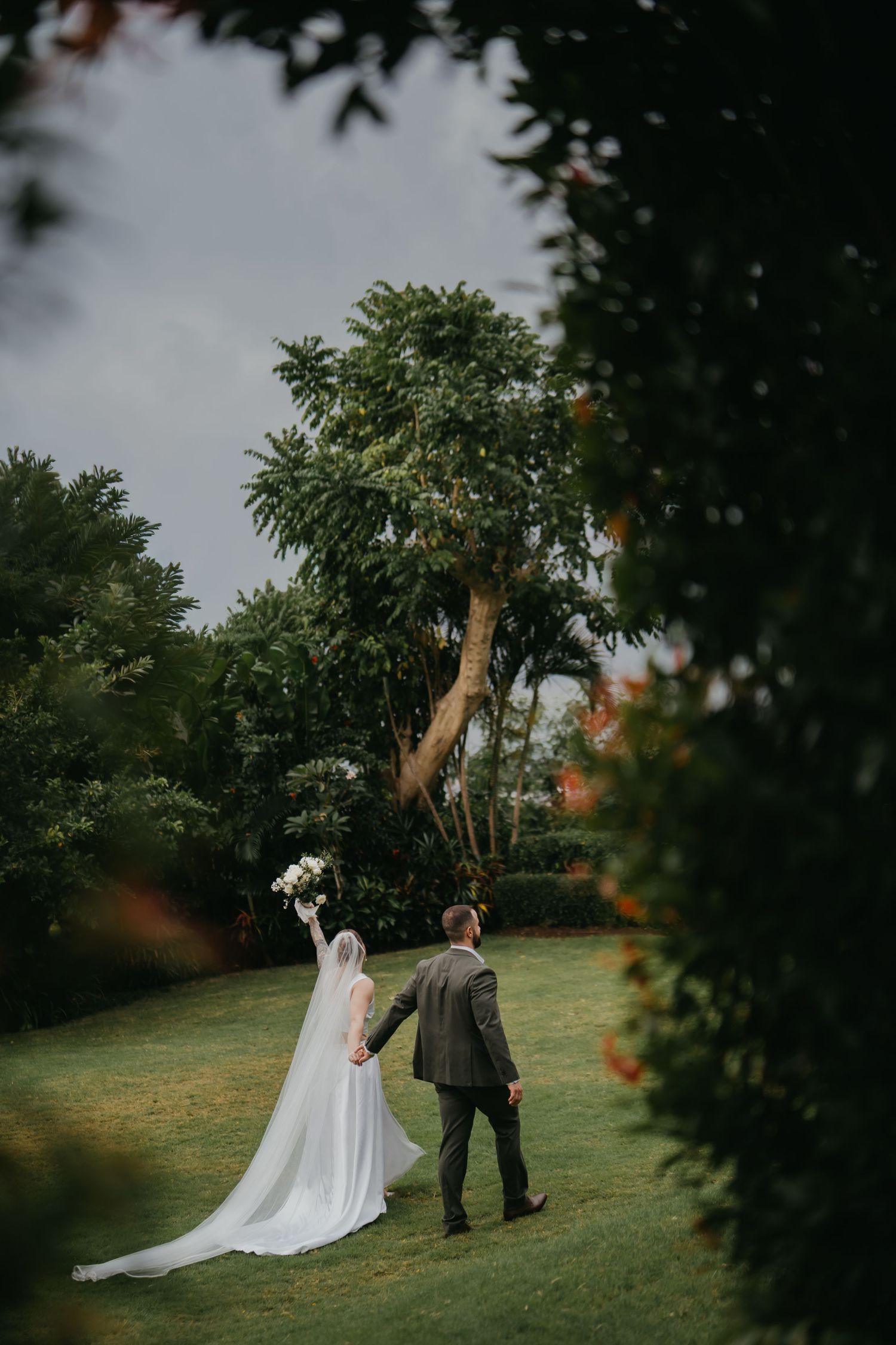 Bride raising bouquet while walking hand in hand with groom through lush green garden setting.