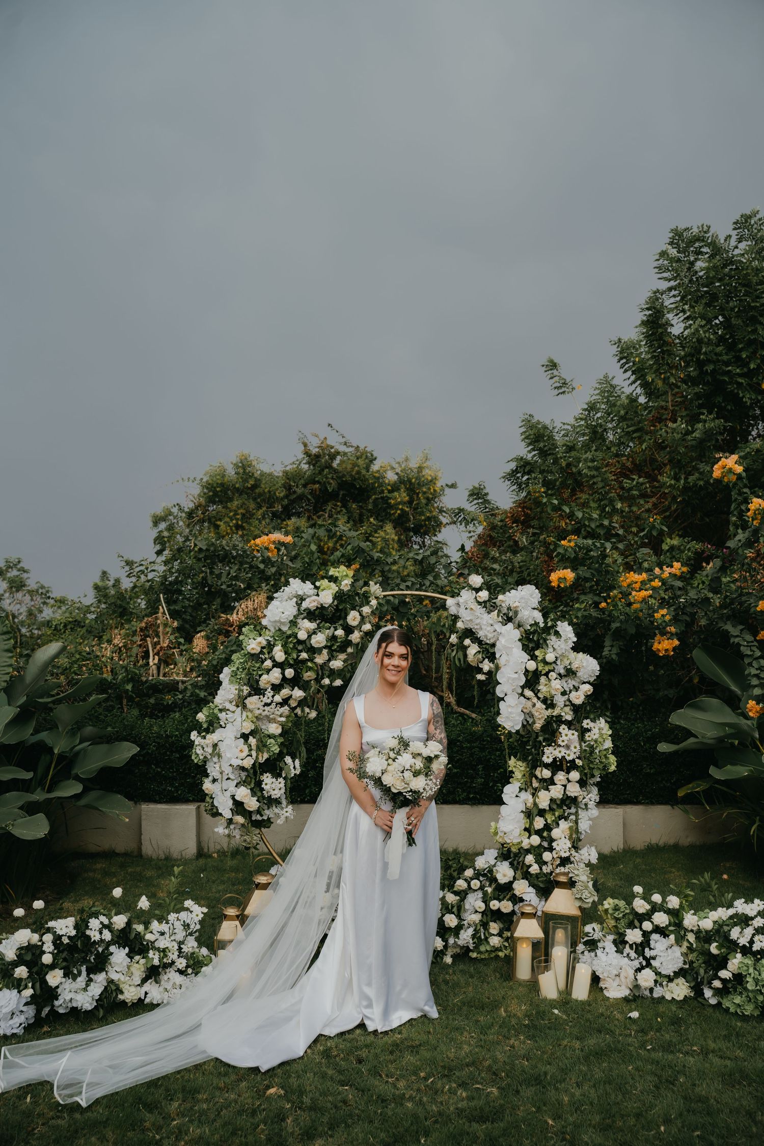 Bride in white gown with veil holding bouquet before circular white floral ceremony arch.