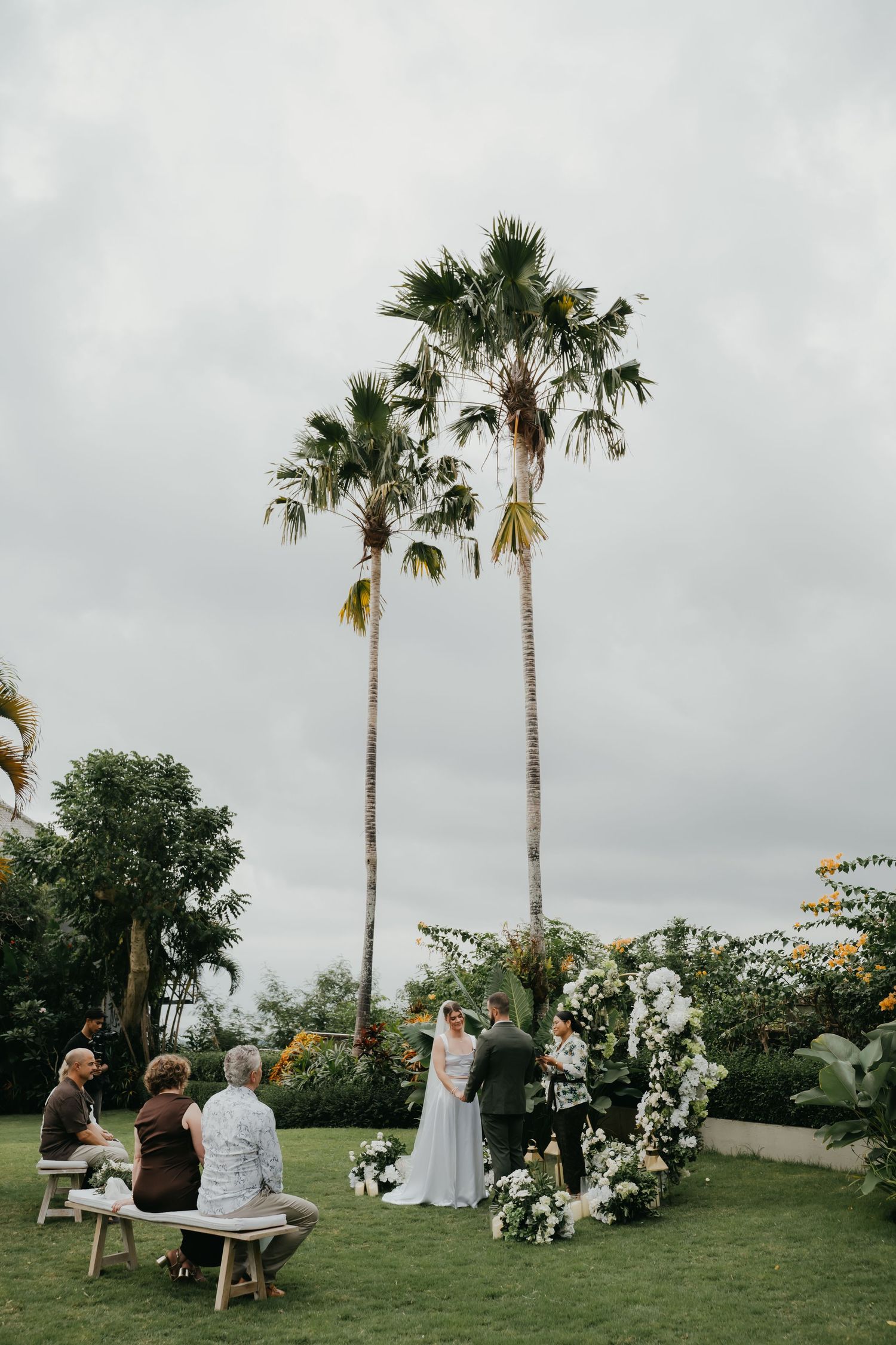 Outdoor garden wedding ceremony with couple at floral arch, guests seated on benches, tall palm trees against cloudy sky.