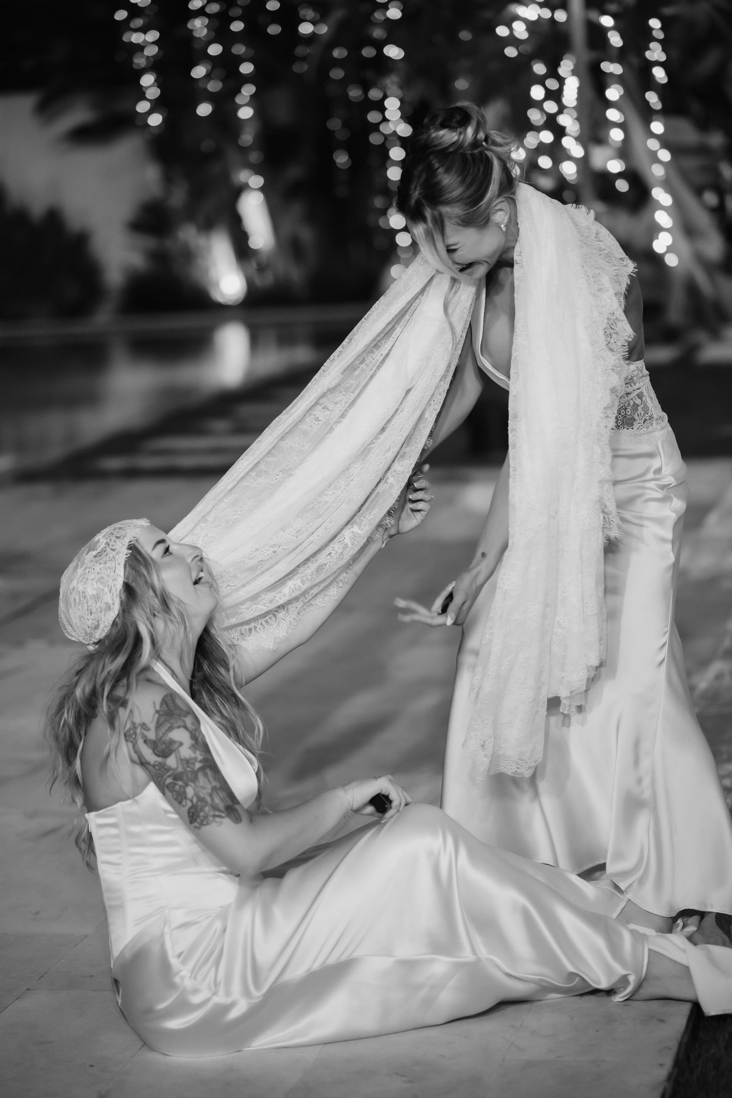 Two brides in white satin gowns share a playful moment with a veil at an outdoor evening wedding reception.