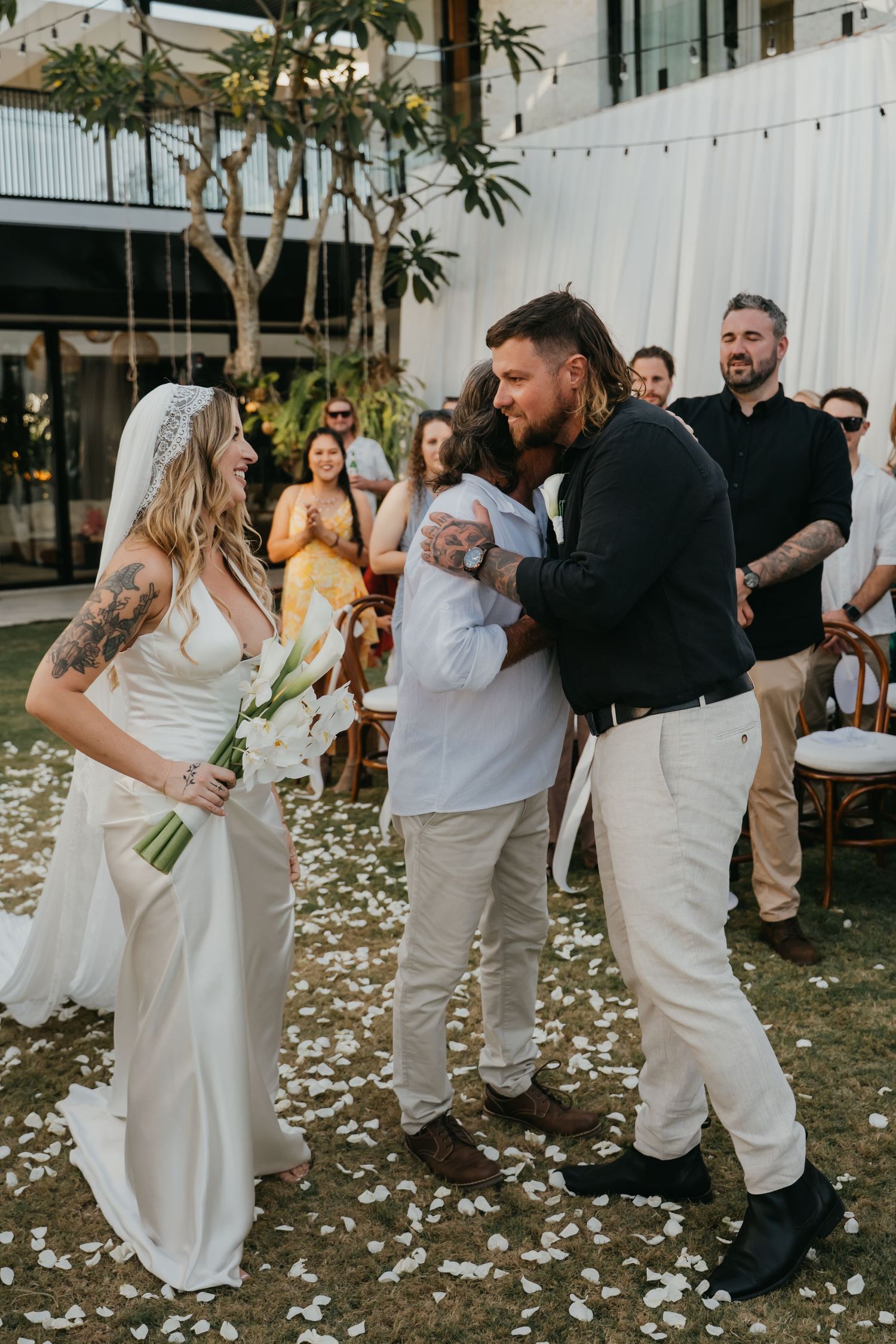 Bride laughing as groom hugs a guest after outdoor wedding ceremony with flower petals on ground.