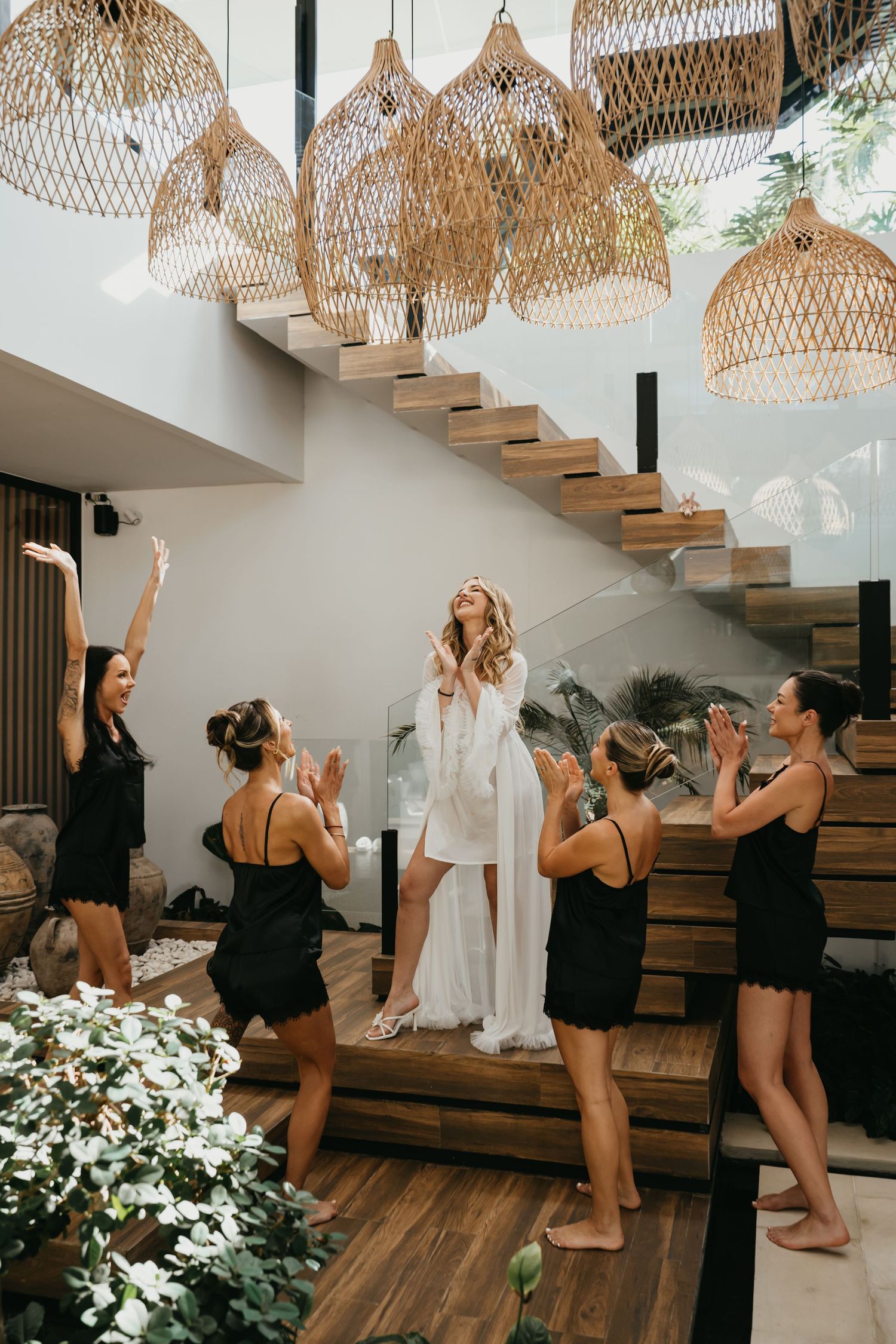 Bride in white robe celebrated by bridesmaids in black on modern staircase with wicker pendant lights.