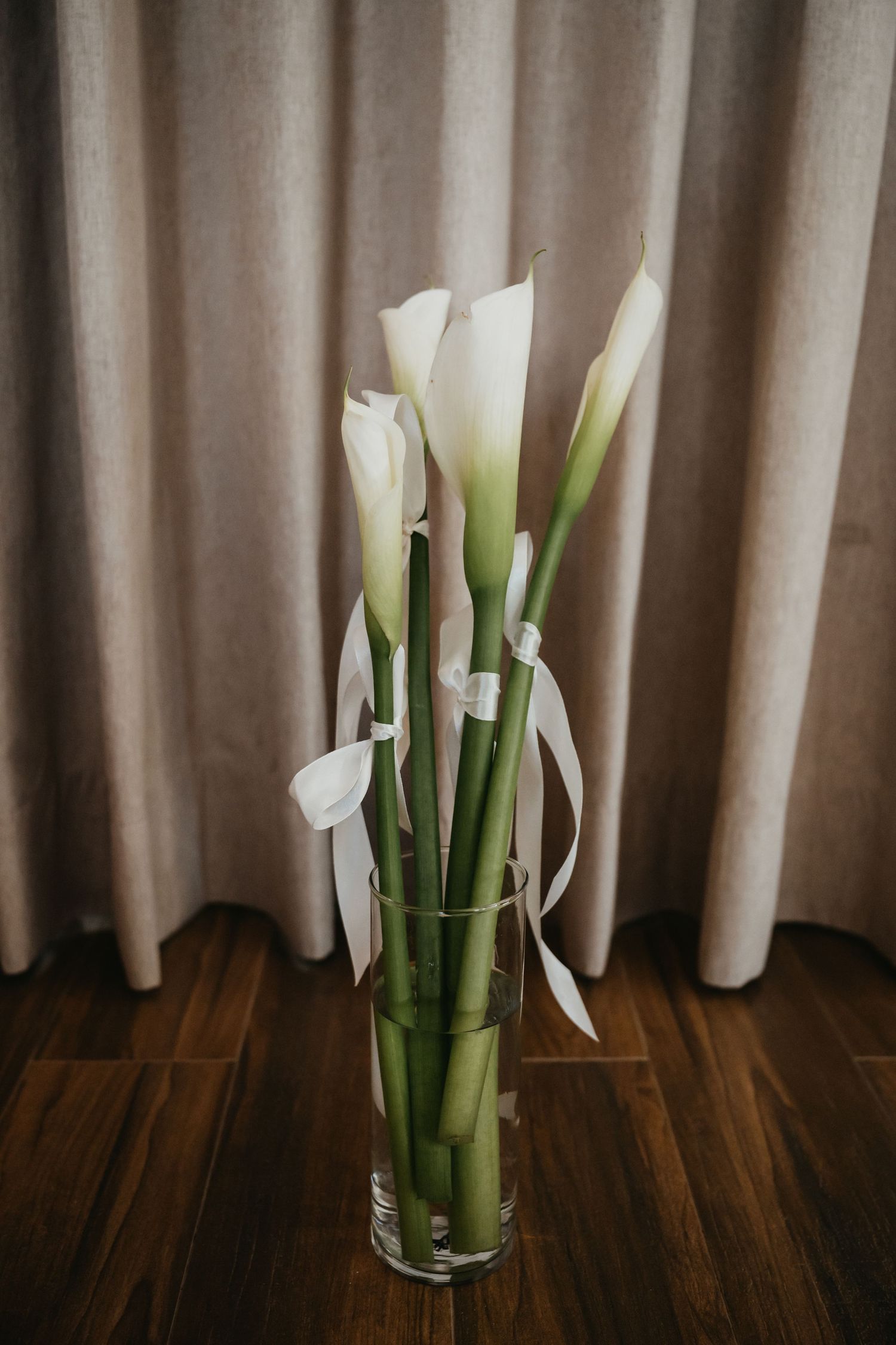 White calla lilies in a glass vase tied with ribbon on hardwood floor.
