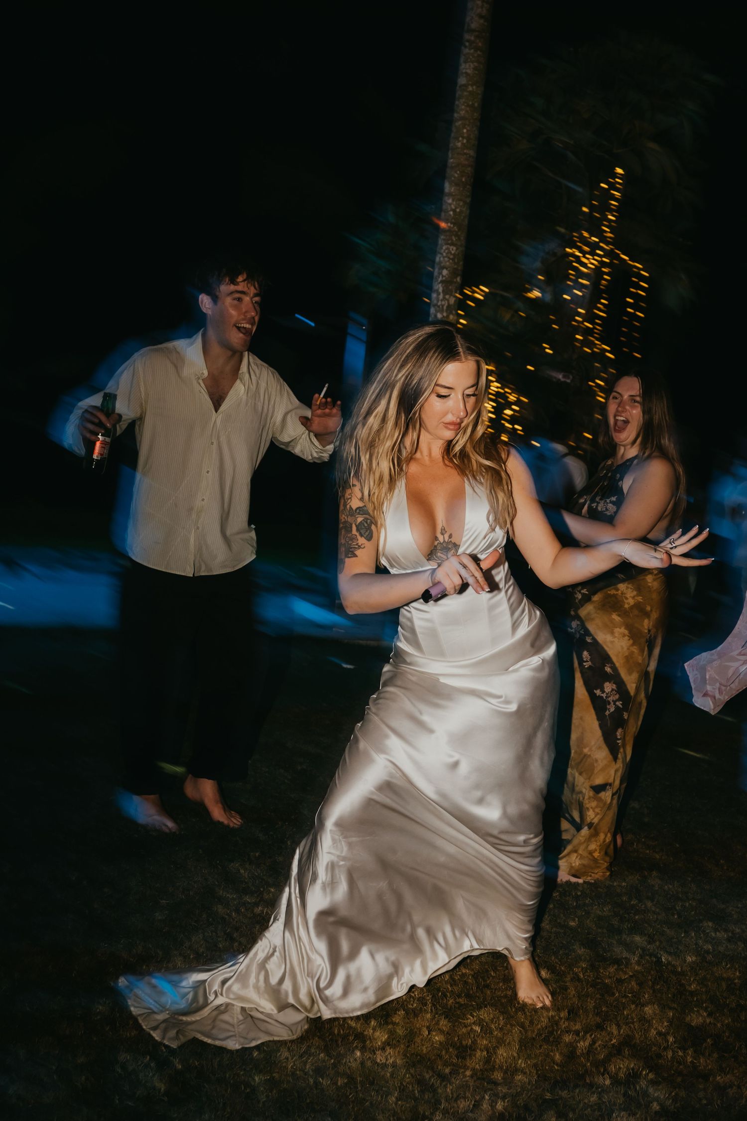 Bride in satin gown dances barefoot with guests at lively outdoor wedding reception under the night sky.