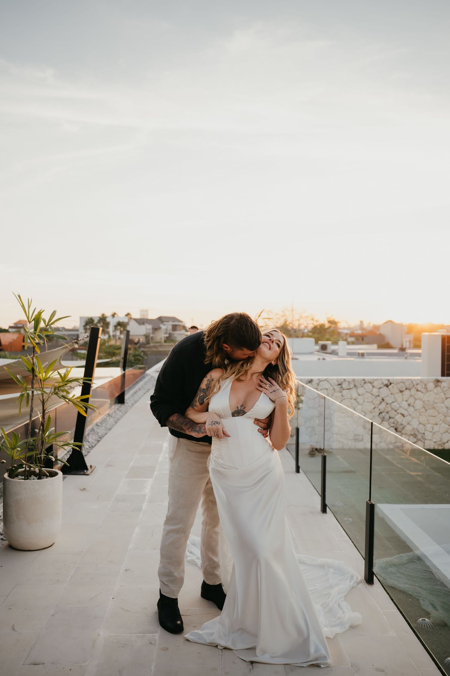 Tattooed groom embracing laughing bride in white gown on rooftop at golden hour sunset.