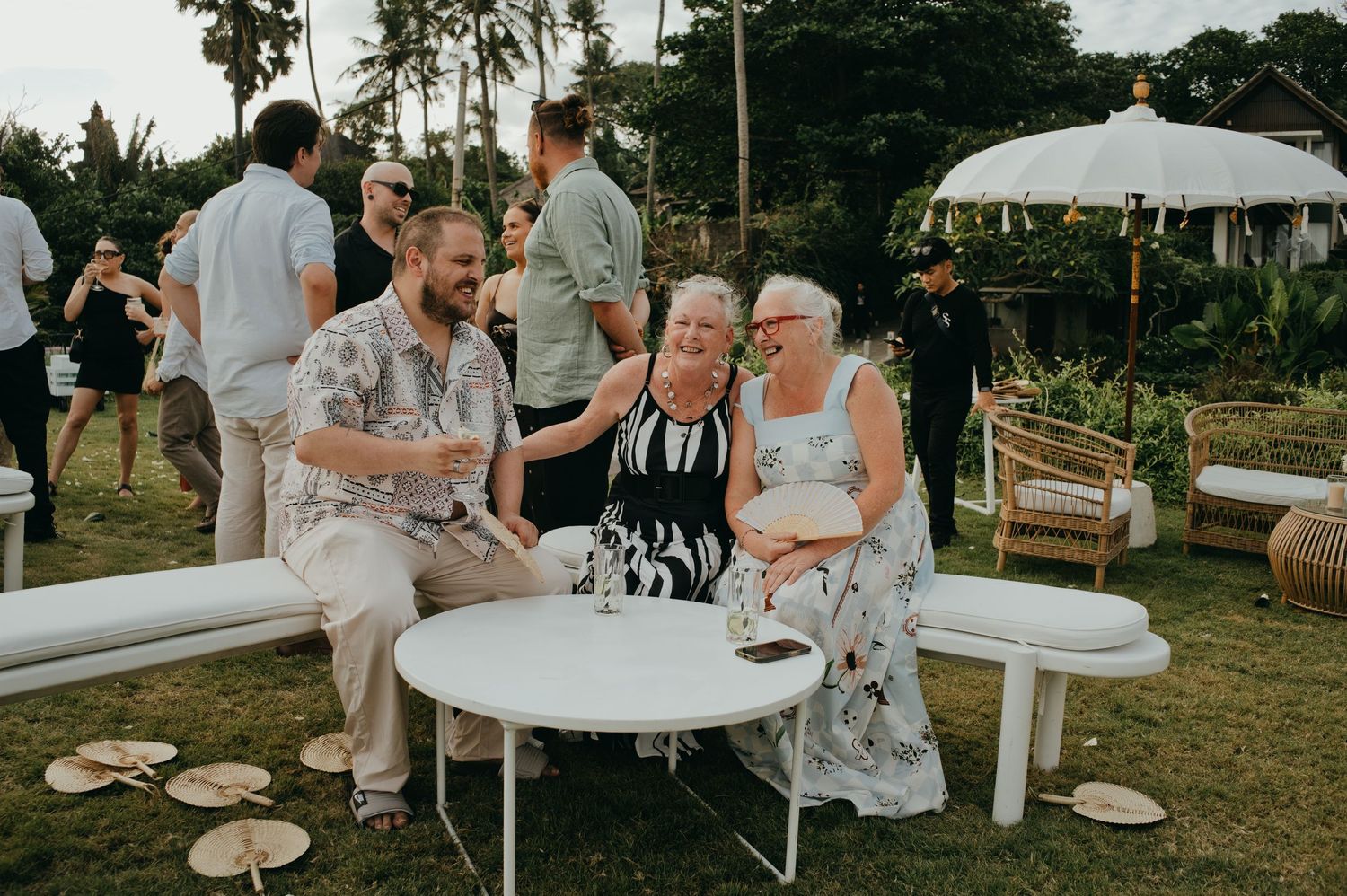 Three guests laughing and socializing at an outdoor tropical wedding reception seating area.