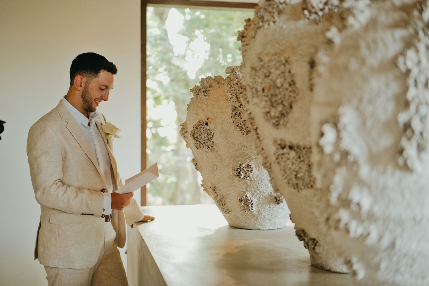 Groom in a beige suit reads vows beside an ornate floral wall sculpture at a modern wedding venue.