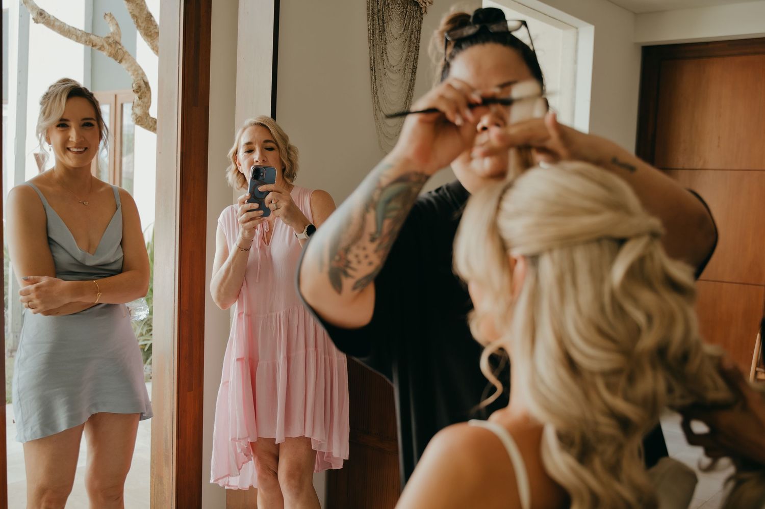 Bride getting hair done while bridesmaids watch and photograph the wedding morning preparations.